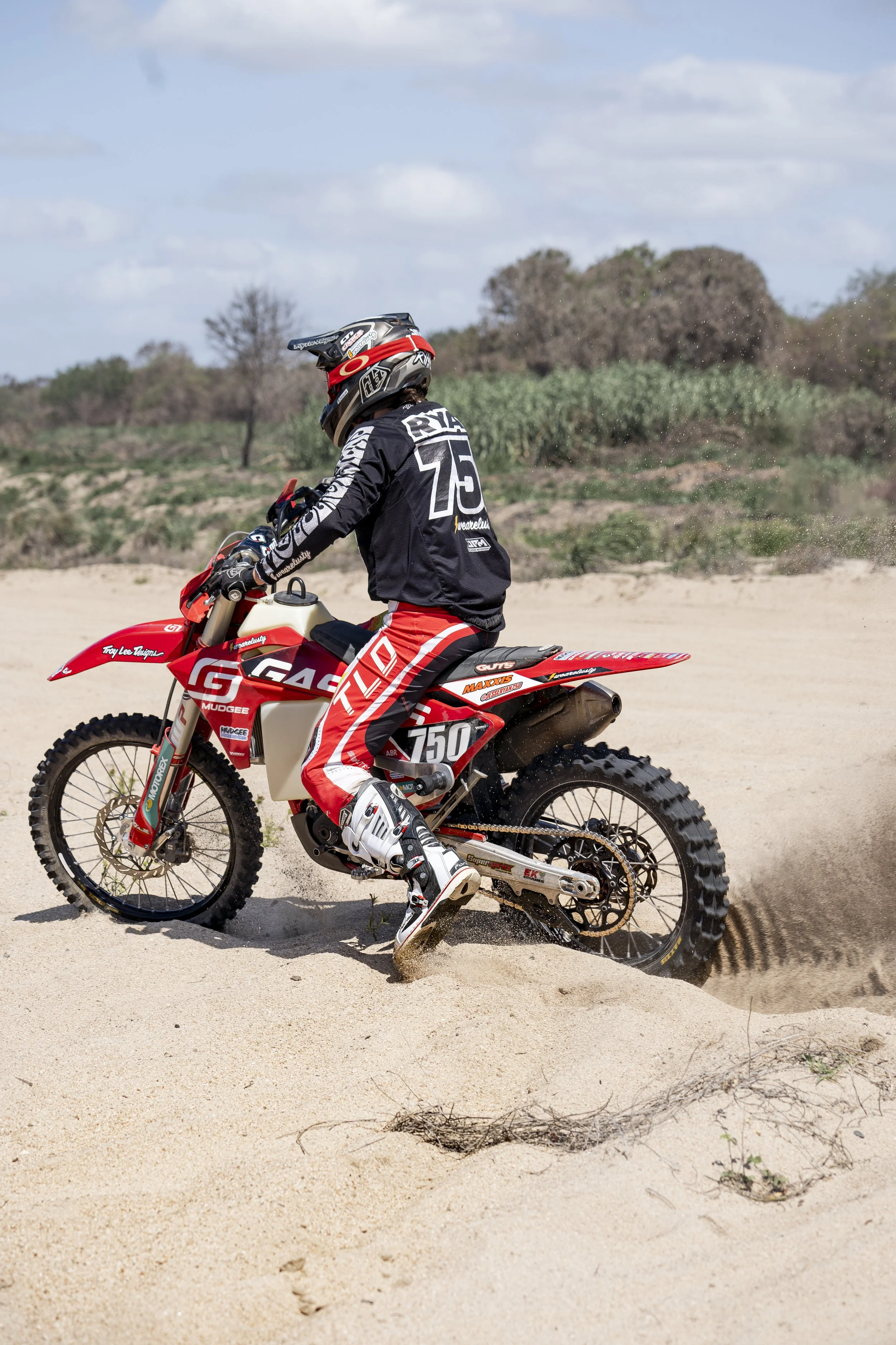 A person riding a red and white dirt bike on a sandy terrain with sparse vegetation and a cloudy sky.