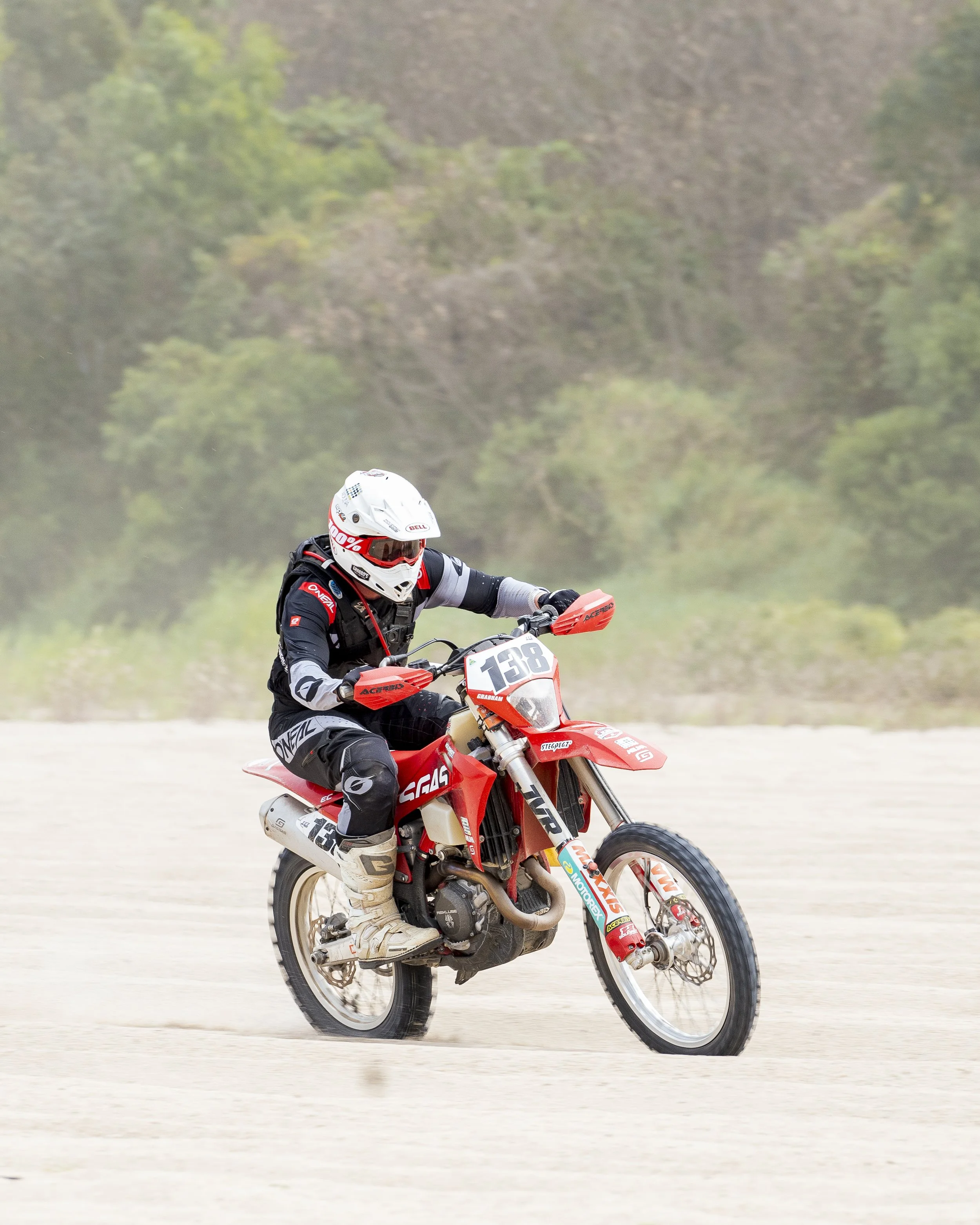 A motocross rider riding a red and white dirt bike on a sandy track, wearing a white helmet, black gear with red and white accents, and beige boots