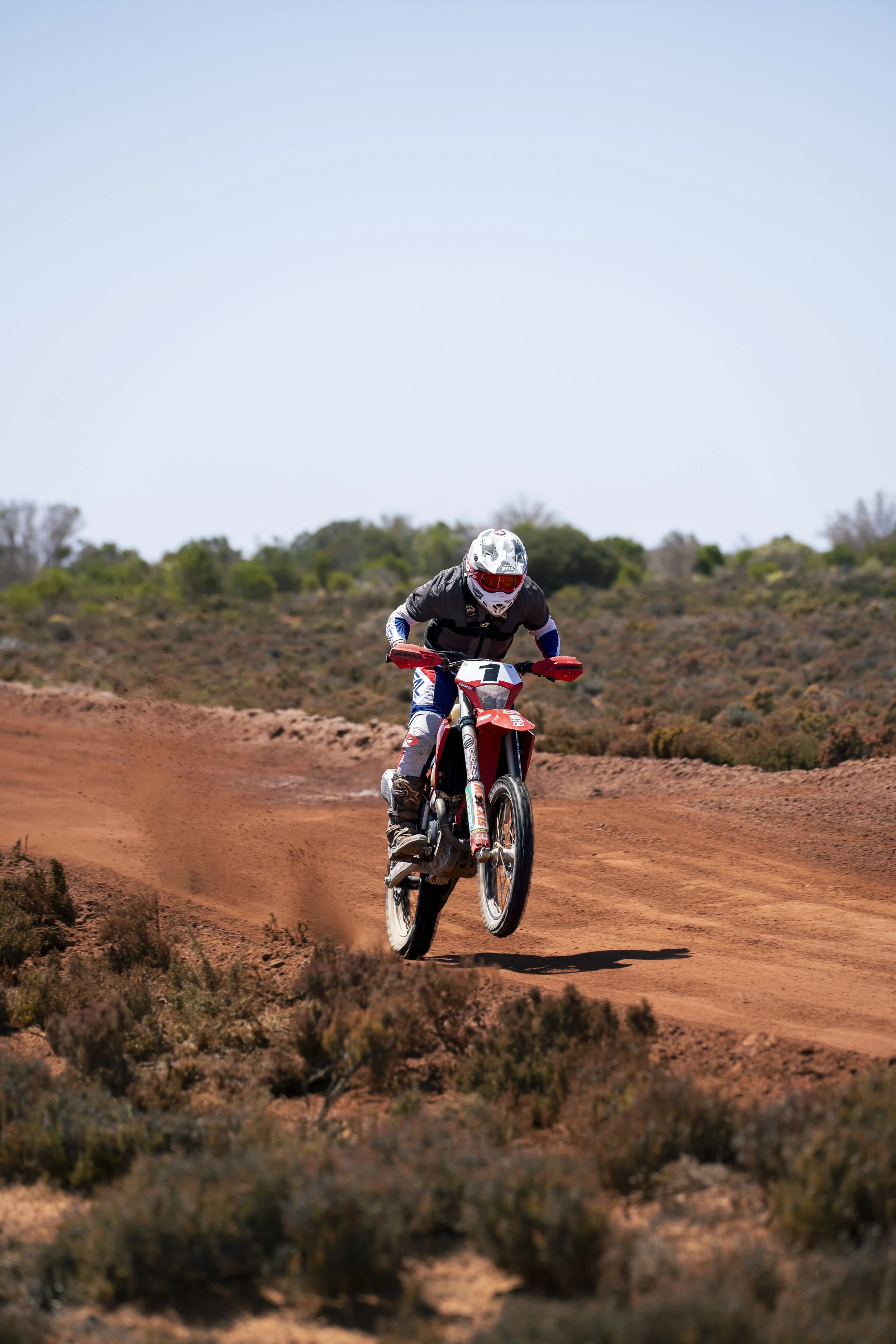 A person riding a motocross dirt bike on a dirt trail in a desert-like landscape with bushes and sparse trees under a clear blue sky.