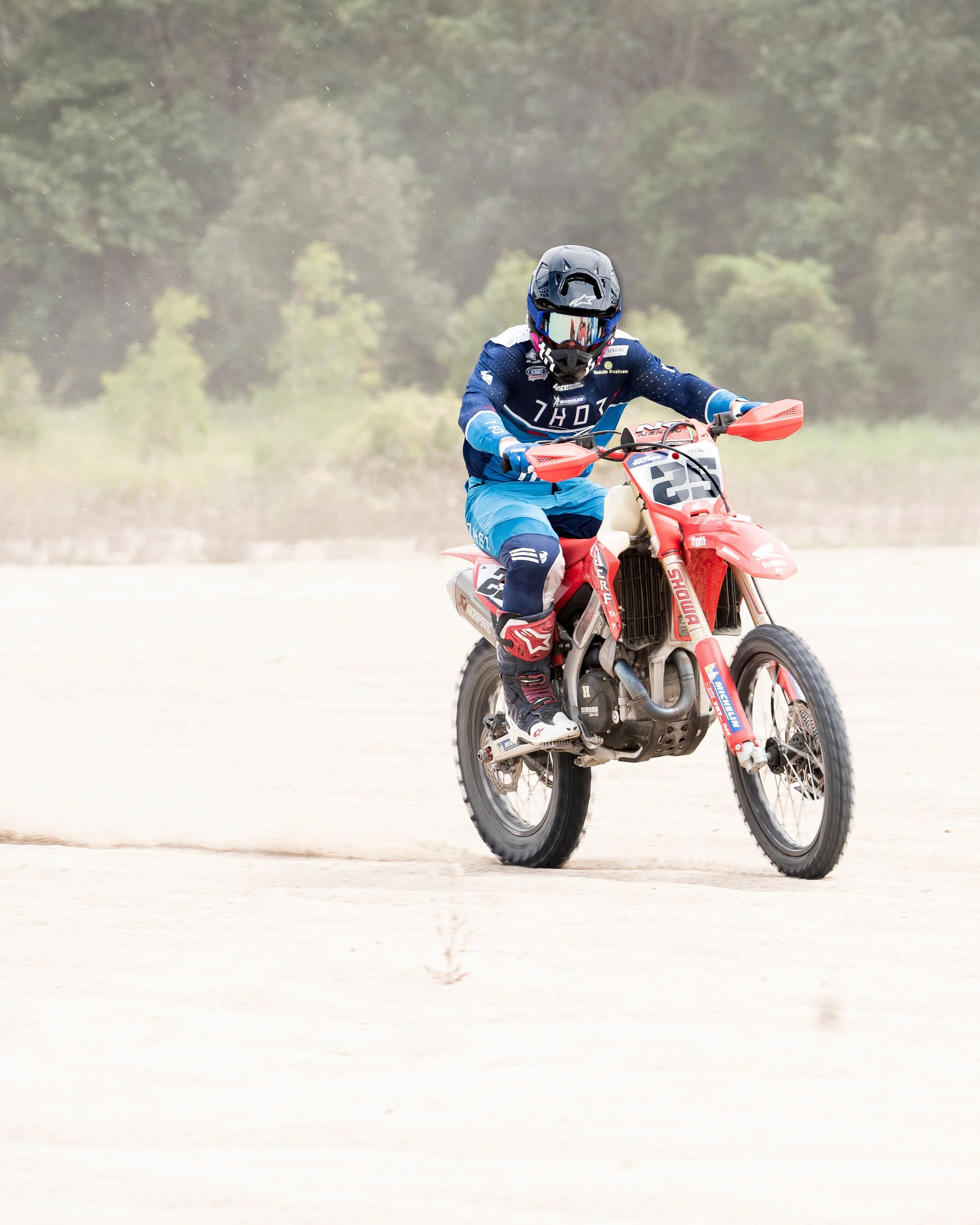 A Motocross rider in blue gear riding a red dirt bike on a sandy trail with trees in the background.