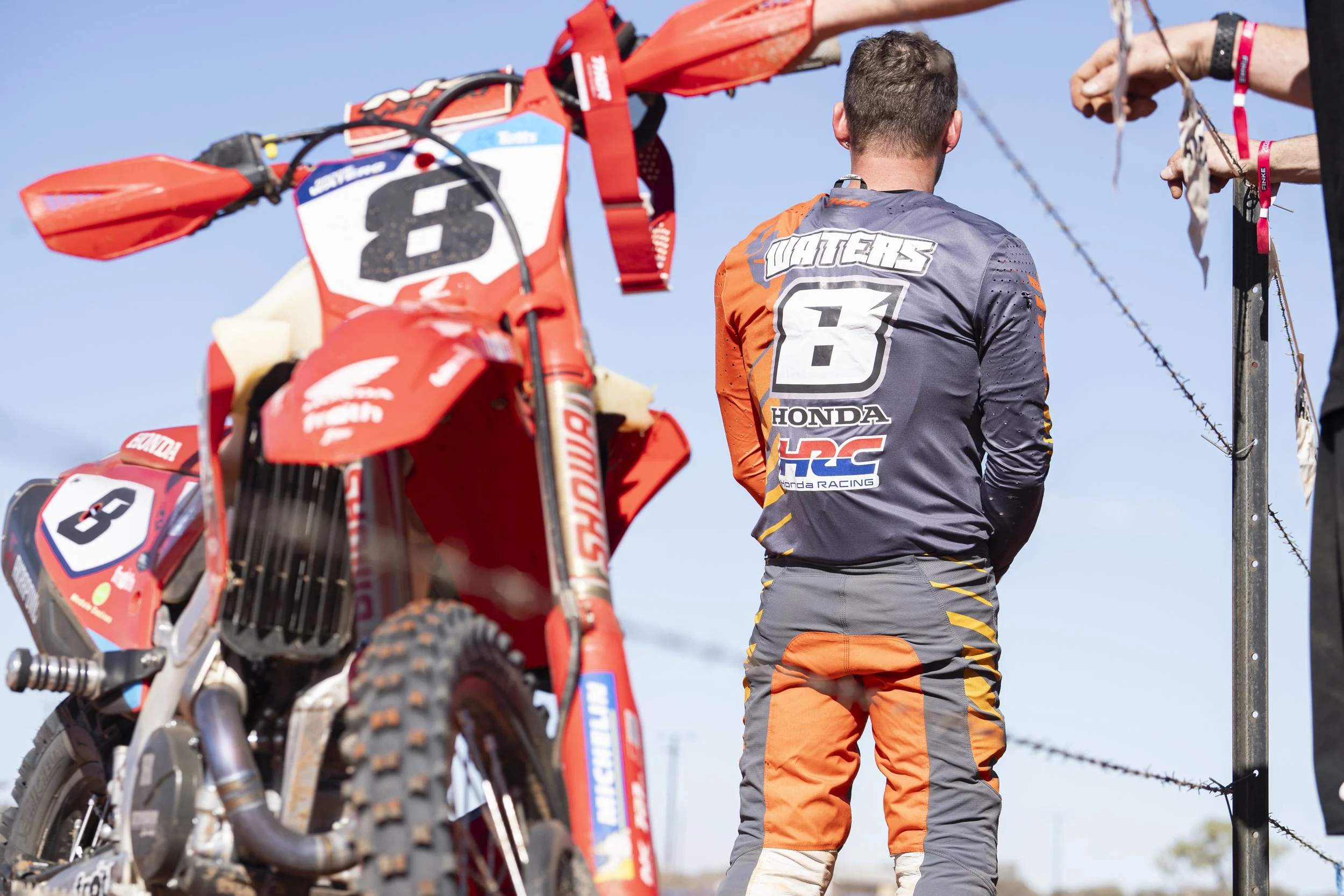 A person in motocross gear stands with their back to the camera, near a red Honda dirt bike, at an outdoor motocross track during daytime with a clear blue sky.