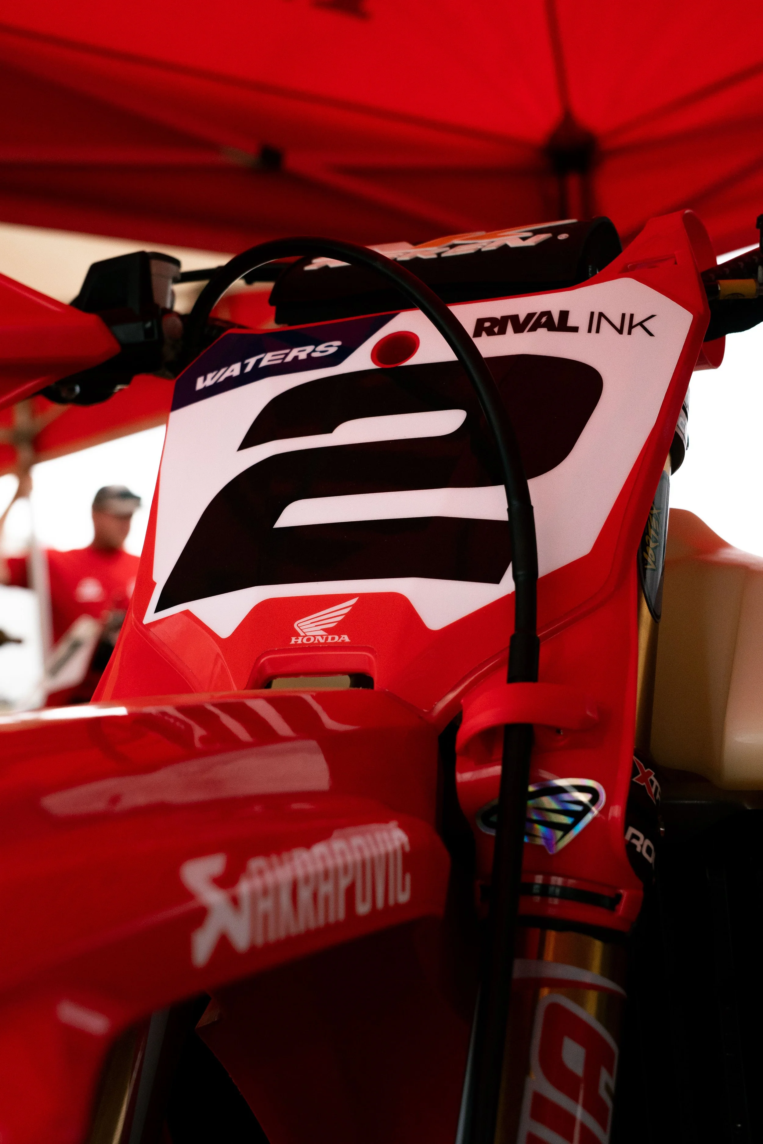 Close-up of a red racing motorcycle with sponsor logos, including Honda and Snap-on, under a red canopy with a blurred person in the background.