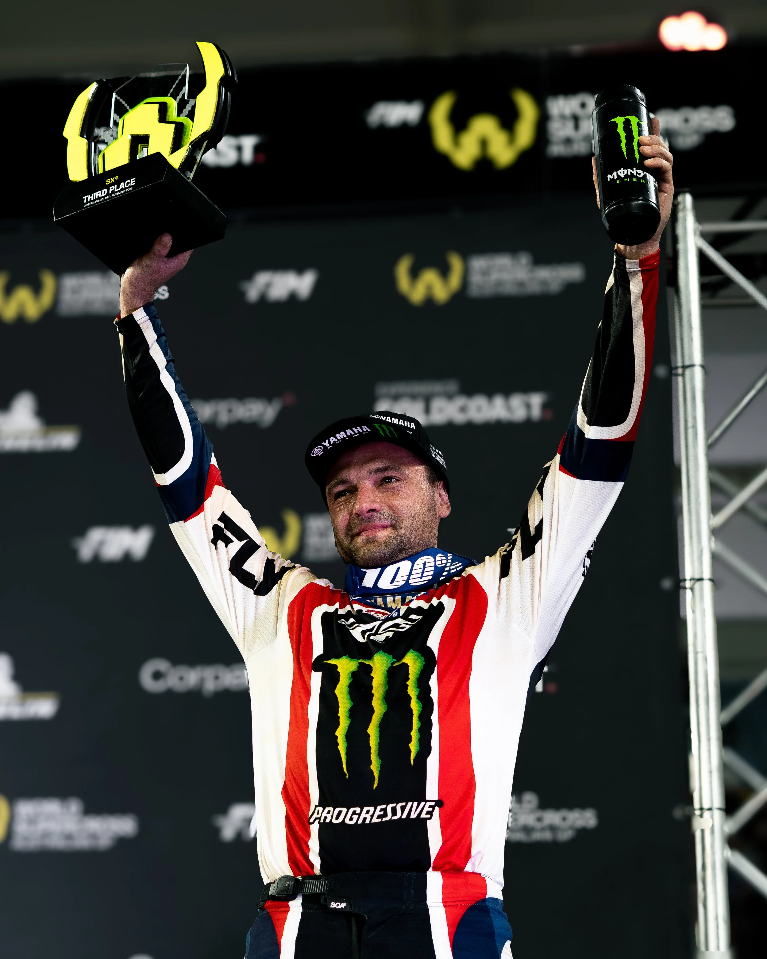 A race car driver in racing gear holding a trophy and a drink, celebrating on the podium after a race.