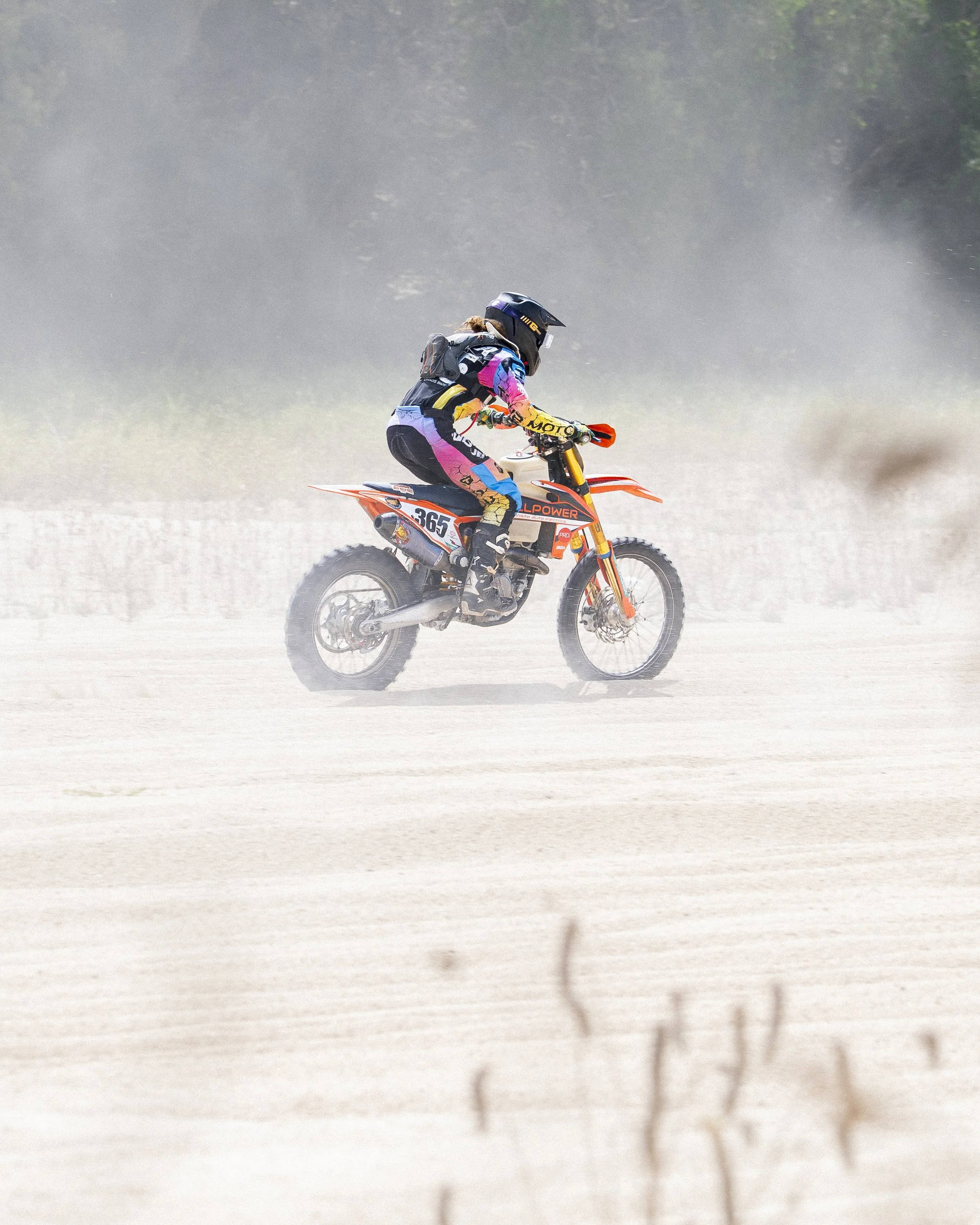 A person riding a dirt bike on a dusty trail, wearing colorful gear and a helmet, with a blurred background of trees and dust.
