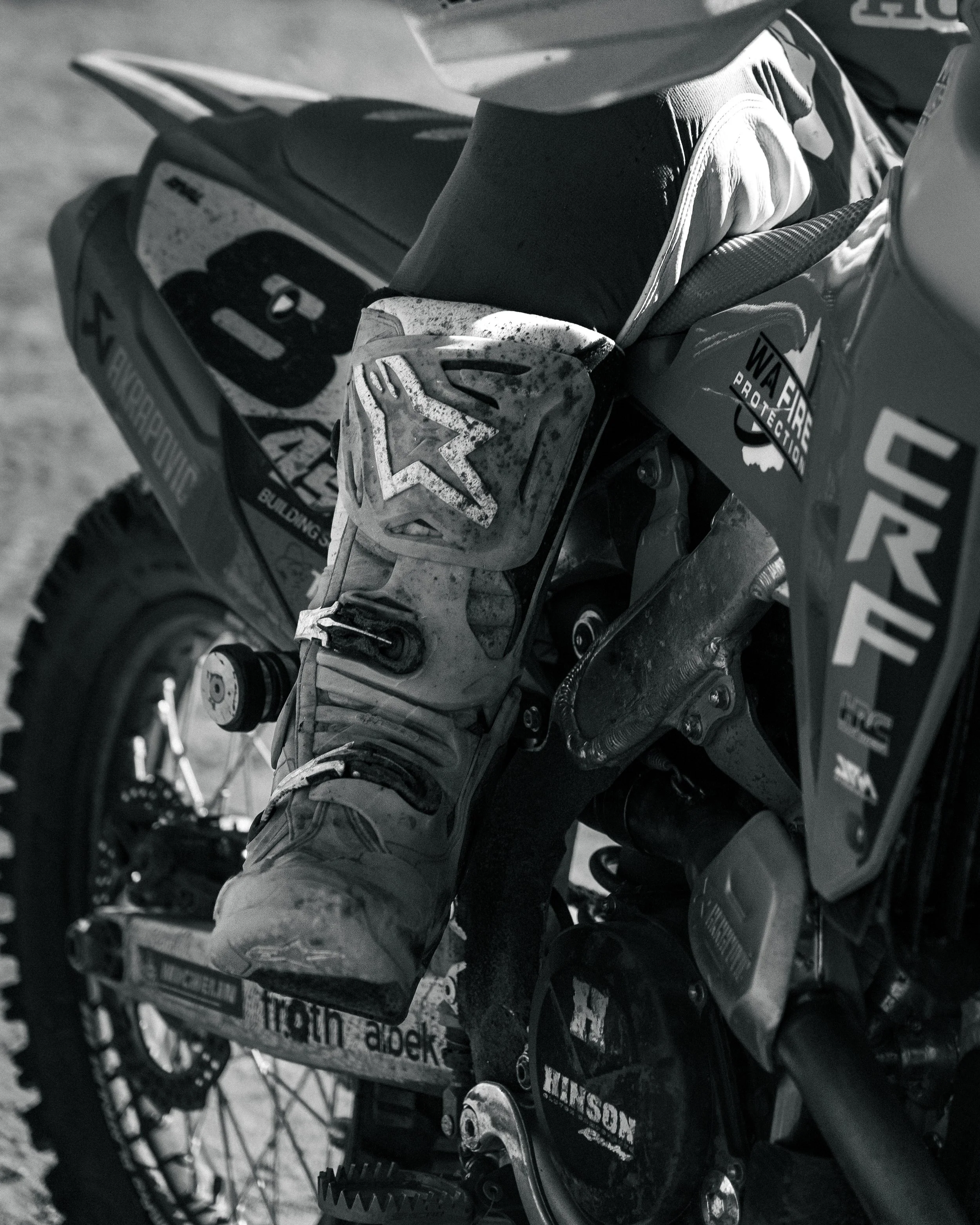 Close-up of a dirt bike with a muddy racing boot resting on the footpeg, featuring various sponsor logos and dirt stains.