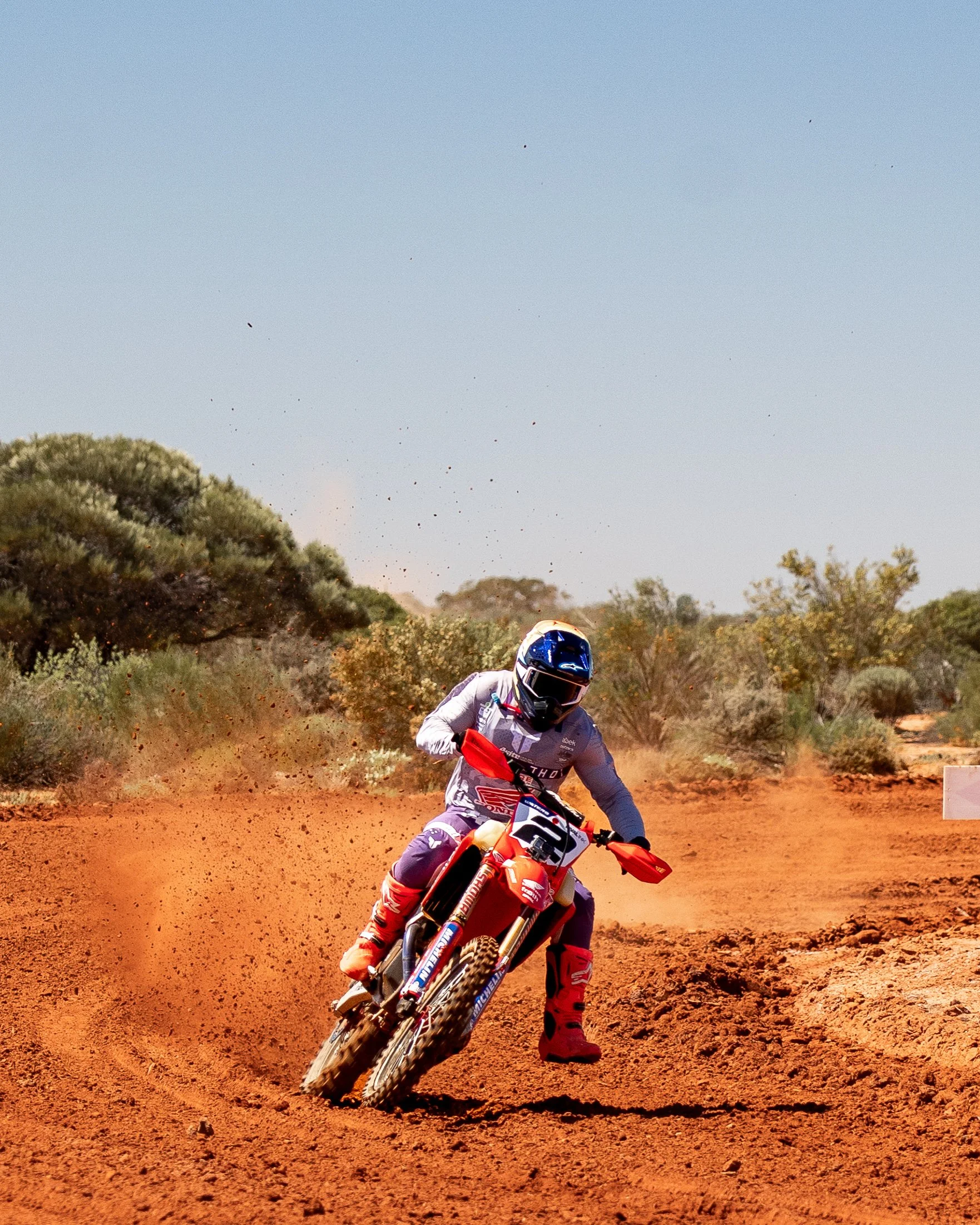 A motocross rider in racing gear and helmet riding a dirt bike on a red dirt track in a desert landscape.