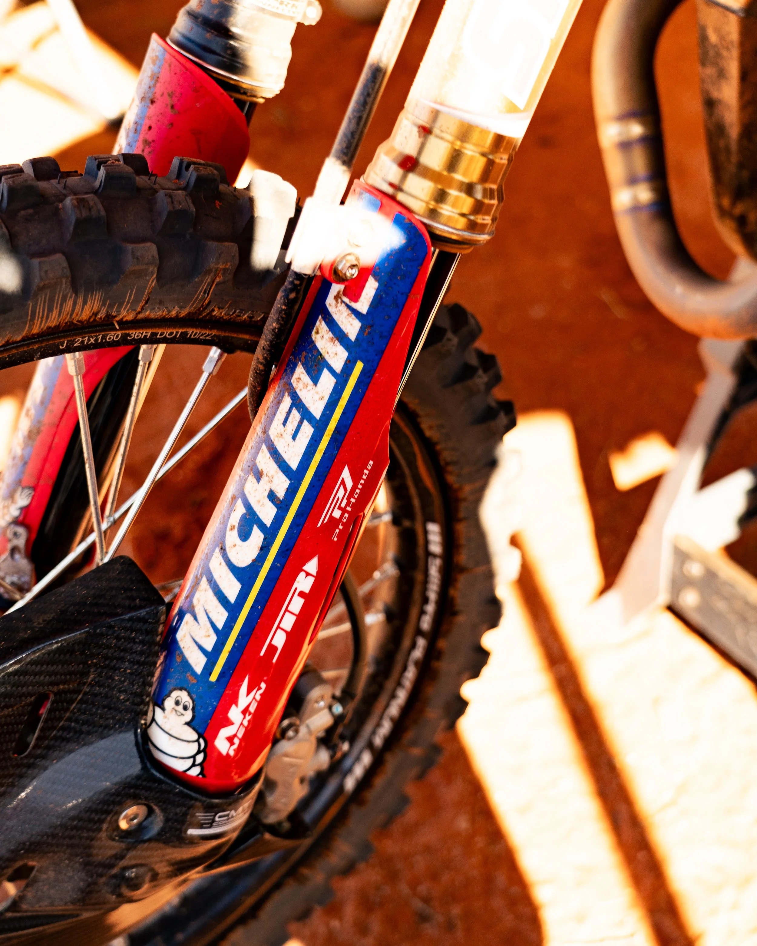 Close-up of a mountain bike front suspension fork with Michelin branding and totem logo, mounted on a trail with reddish dirt.