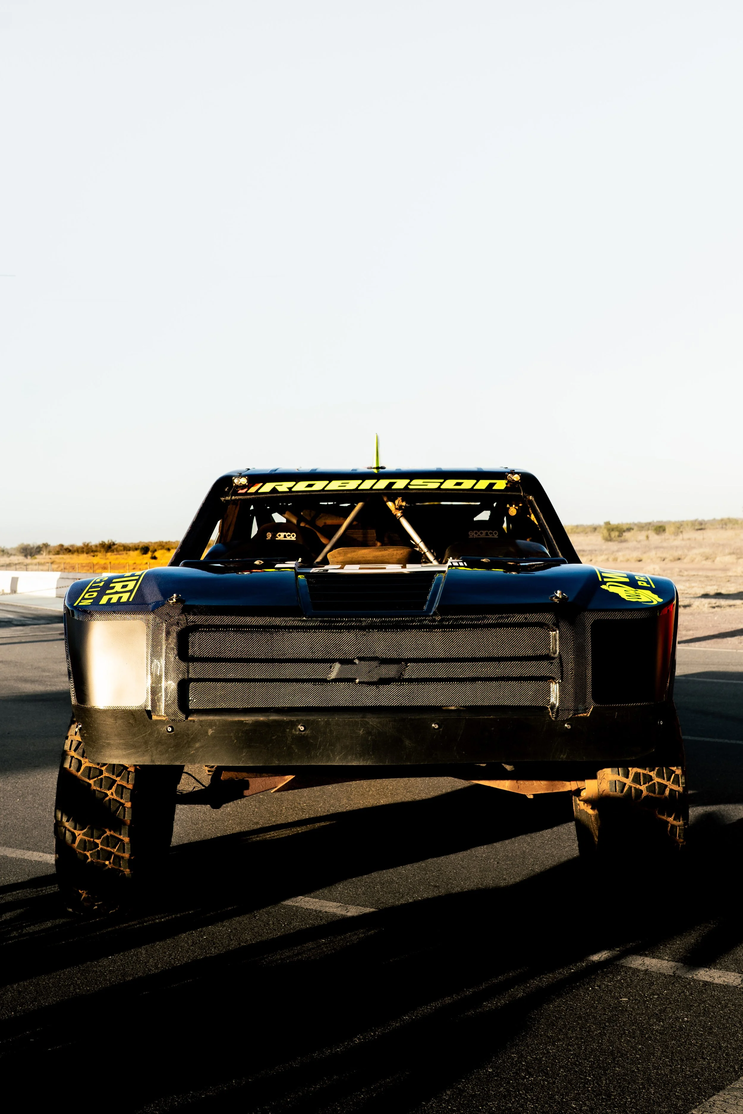 A black off-road racing truck with yellow decals and the word Robbie on the windshield, parked on a race track with a barren desert landscape in the background during daytime.