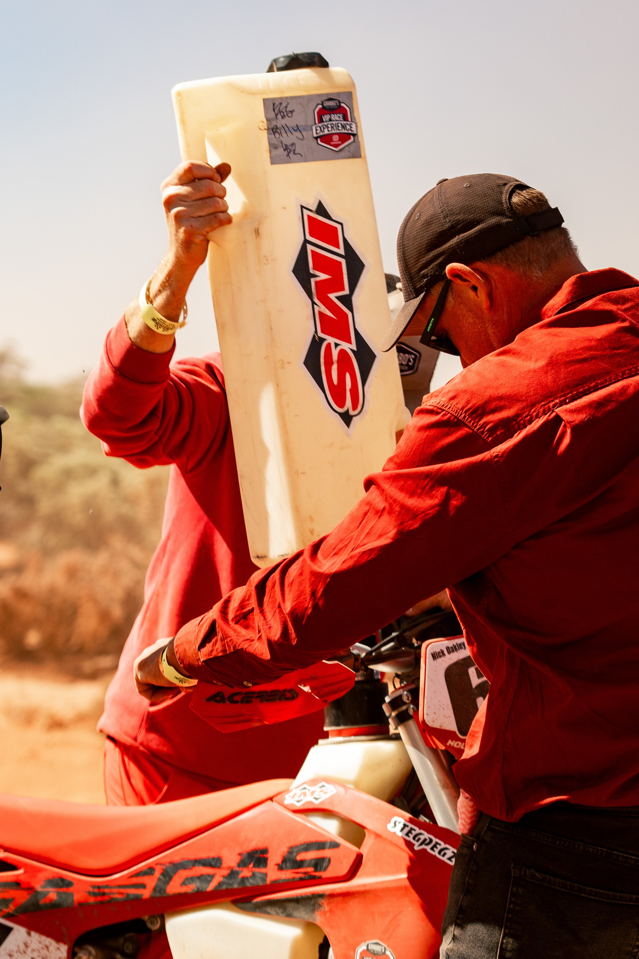 Two men preparing a dirt bike for a race, one holding a large container, both dressed in red and black gear, with a desert landscape in the background.