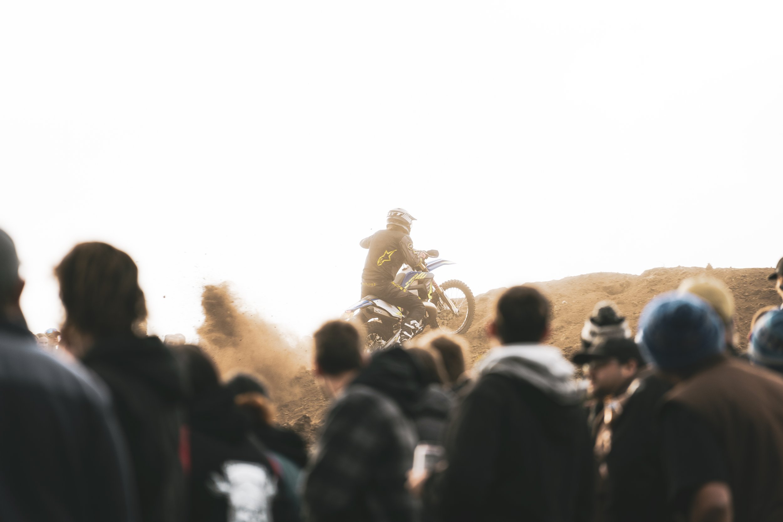 Motocross rider in gear on a dirt track jumping with dust clouds behind him while a crowd of spectators watches.