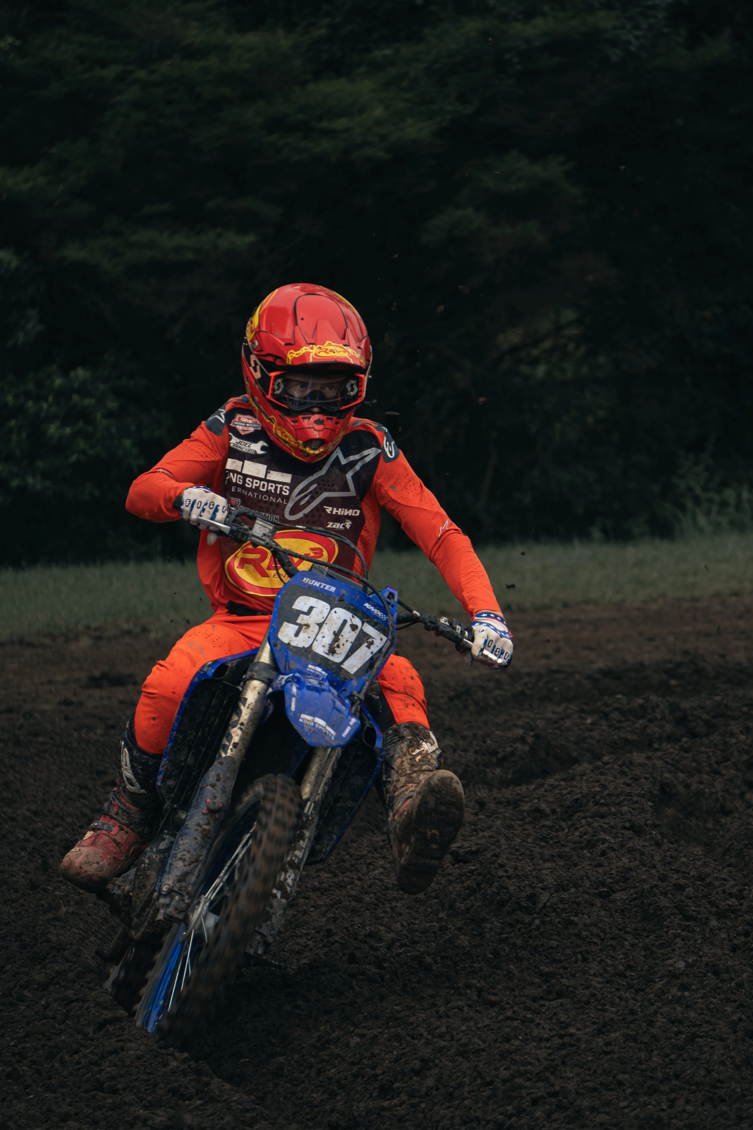 Motorcycle racer in red gear riding on dirt track with mud flying up, number 307 on blue bike, dark wooded background.