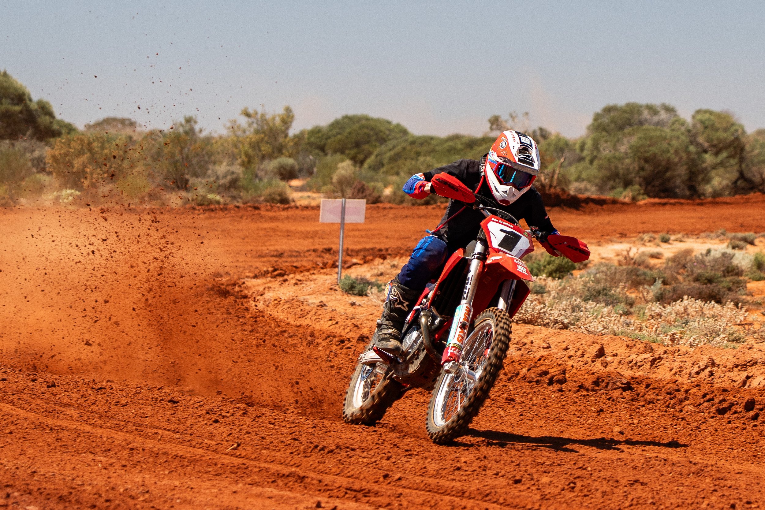 Person riding a dirt bike on a red dirt trail in a desert area with shrubs and sparse trees, wearing a helmet and protective gear
