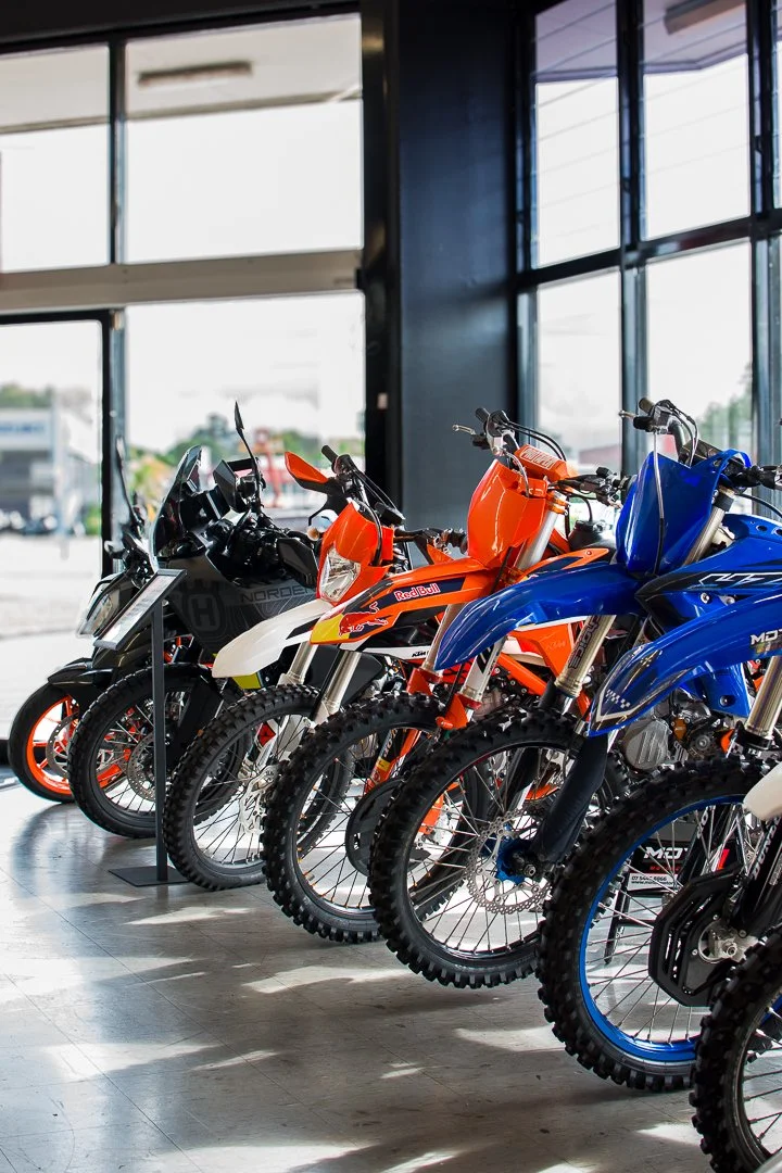 Row of motocross bikes inside a showroom, with large windows in the background.