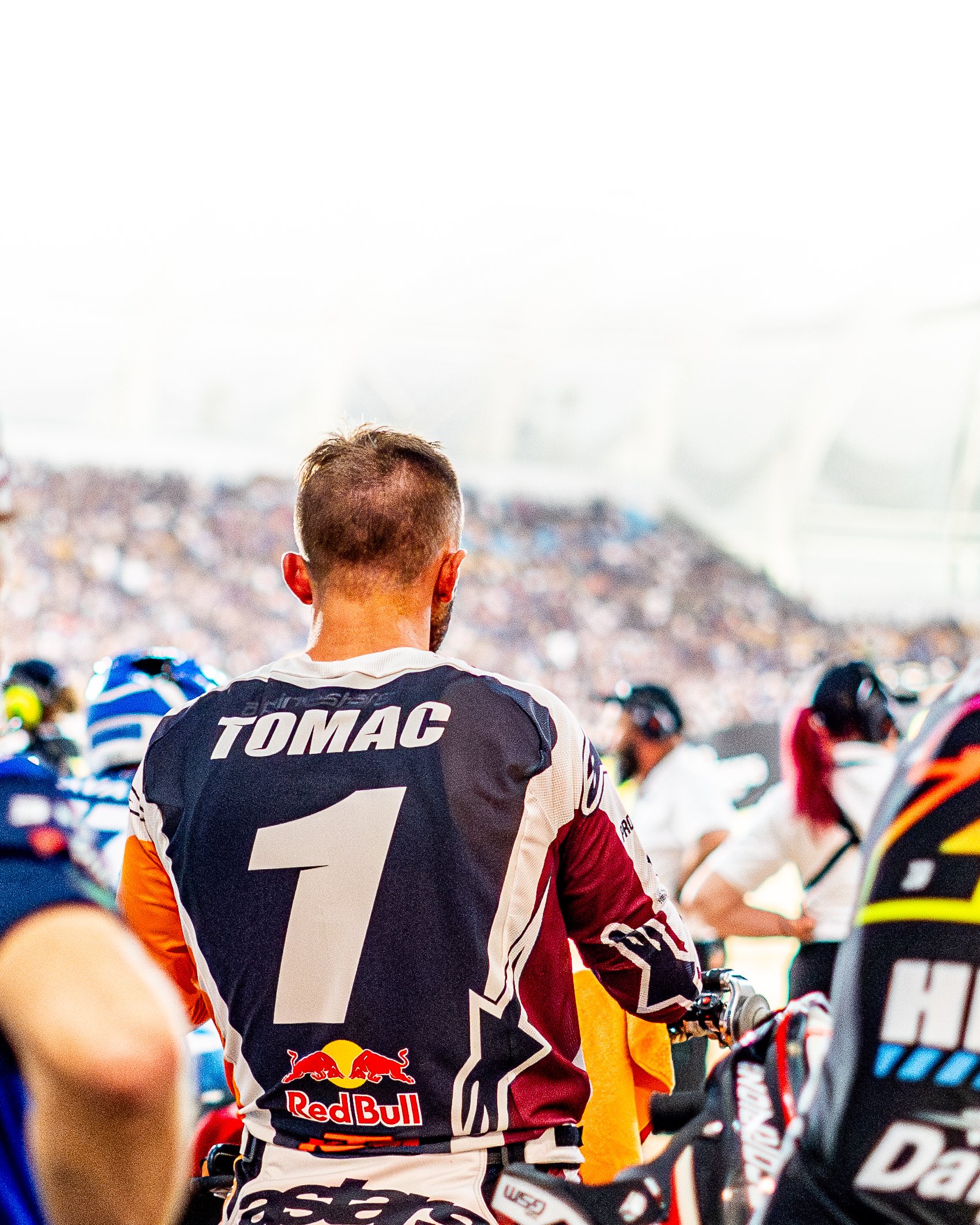 Back view of a motorcycle racer in a racing jersey with the name 'TOMAC' and the number 1 on it, standing in a crowded racing pit area.