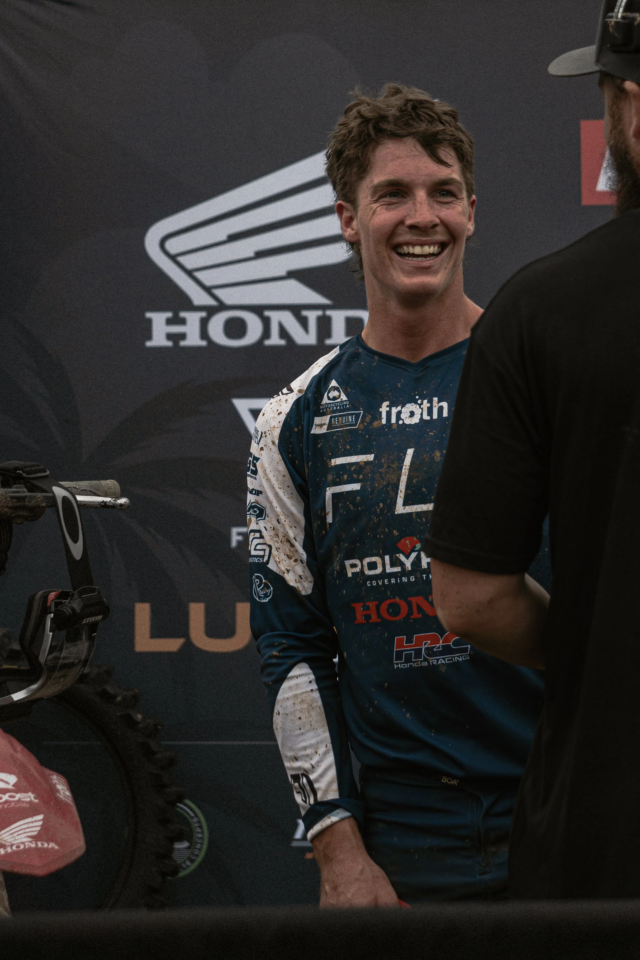 A young man with a big smile, wearing a dark blue motocross jersey with various logos, stands in front of a black backdrop with the Honda logo, talking to another person at a motorsport event.