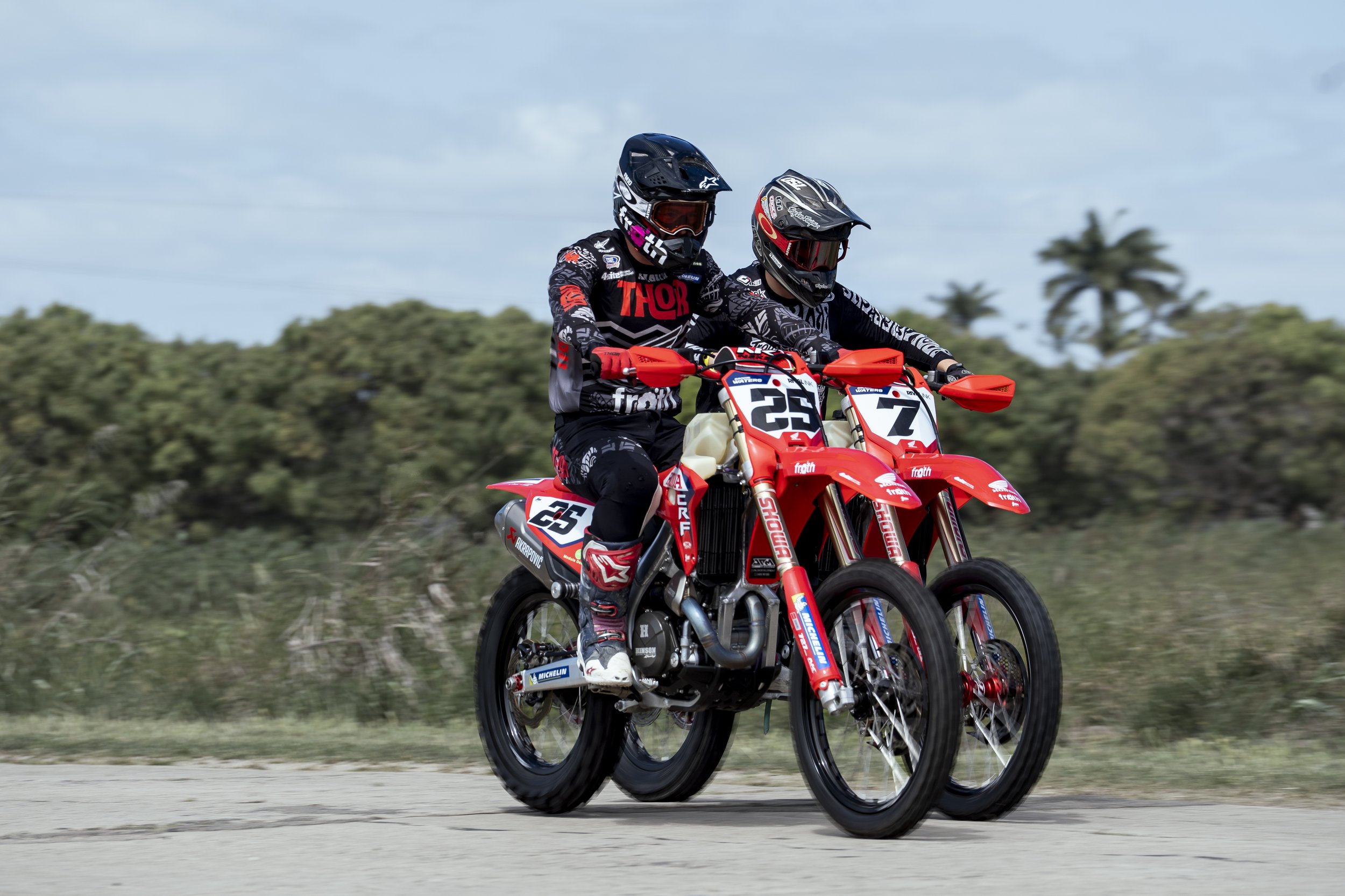 Two dirt bike riders in racing gear riding side by side on an off-road track with trees and cloudy sky in the background.