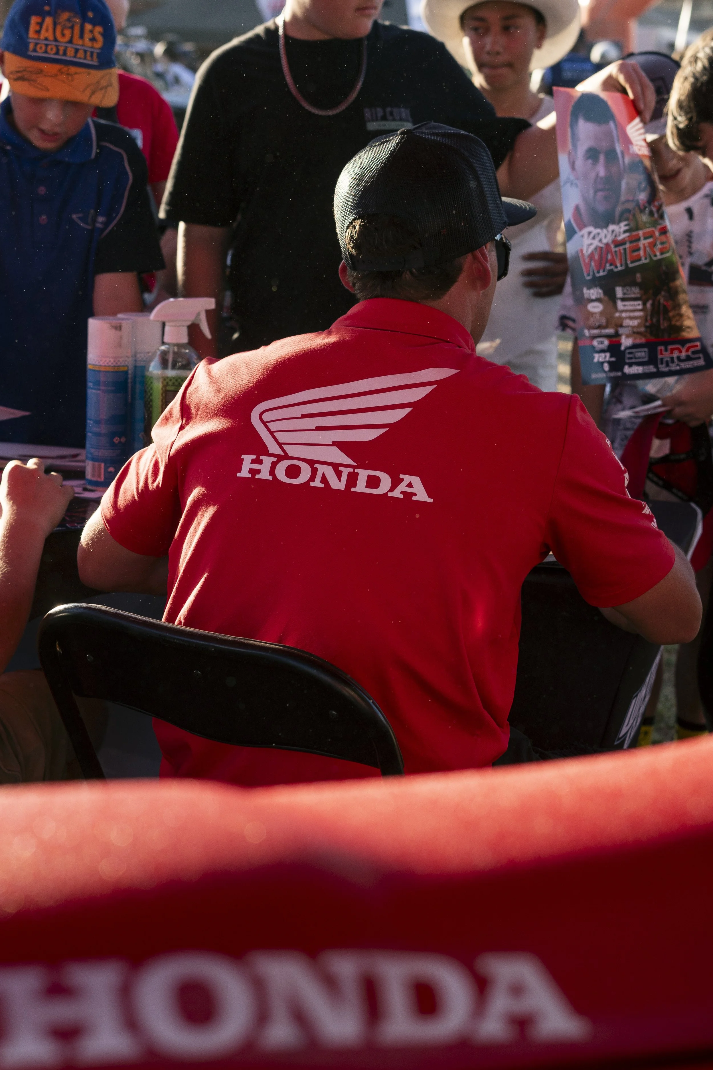 A man wearing a red Honda shirt, black cap, and sunglasses sitting at a table signing autographs for young fans. The scene appears to be at a motorsport event with children holding posters.