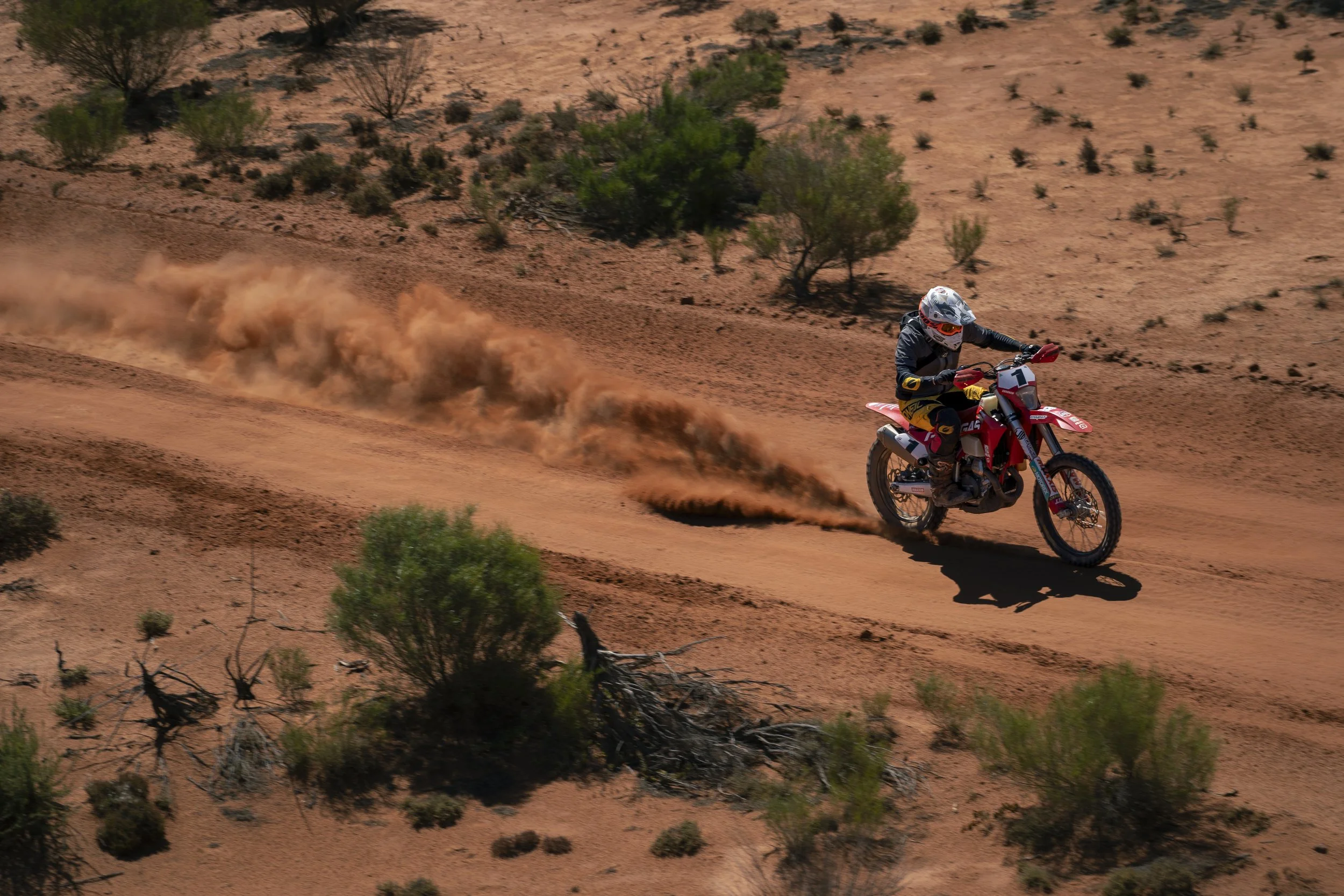 A motocross rider in black gear and a white helmet riding a red dirt bike on a sandy, desert trail, kicking up a trail of dust behind him, with sparse desert vegetation and small bushes around.