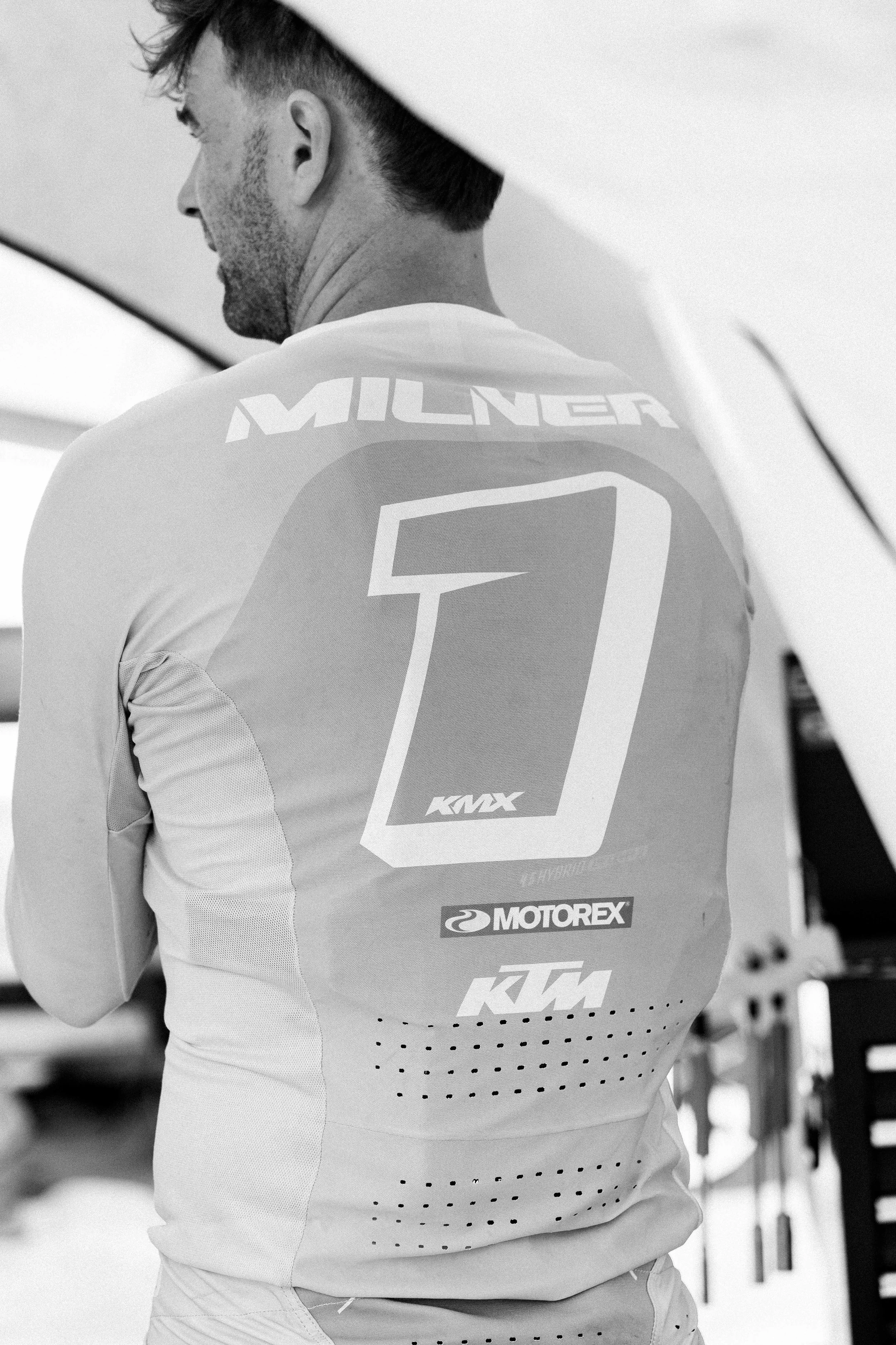 Black and white photo of a man in a racing suit seen from behind, showing large number 1 and various sponsor logos on his back.