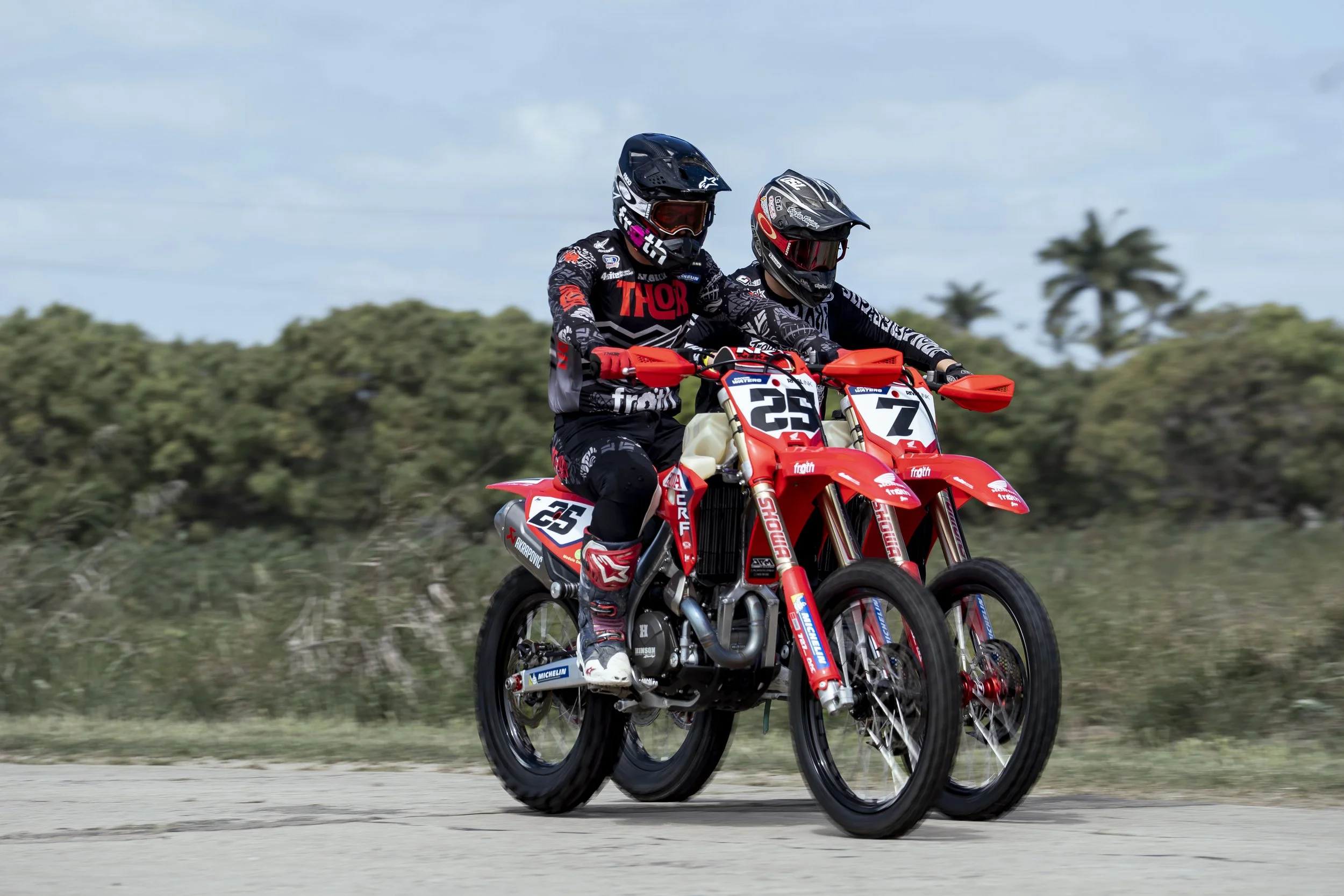 Two motocross riders in racing gear riding orange and white dirt bikes on a dirt track with greenery and trees in the background.