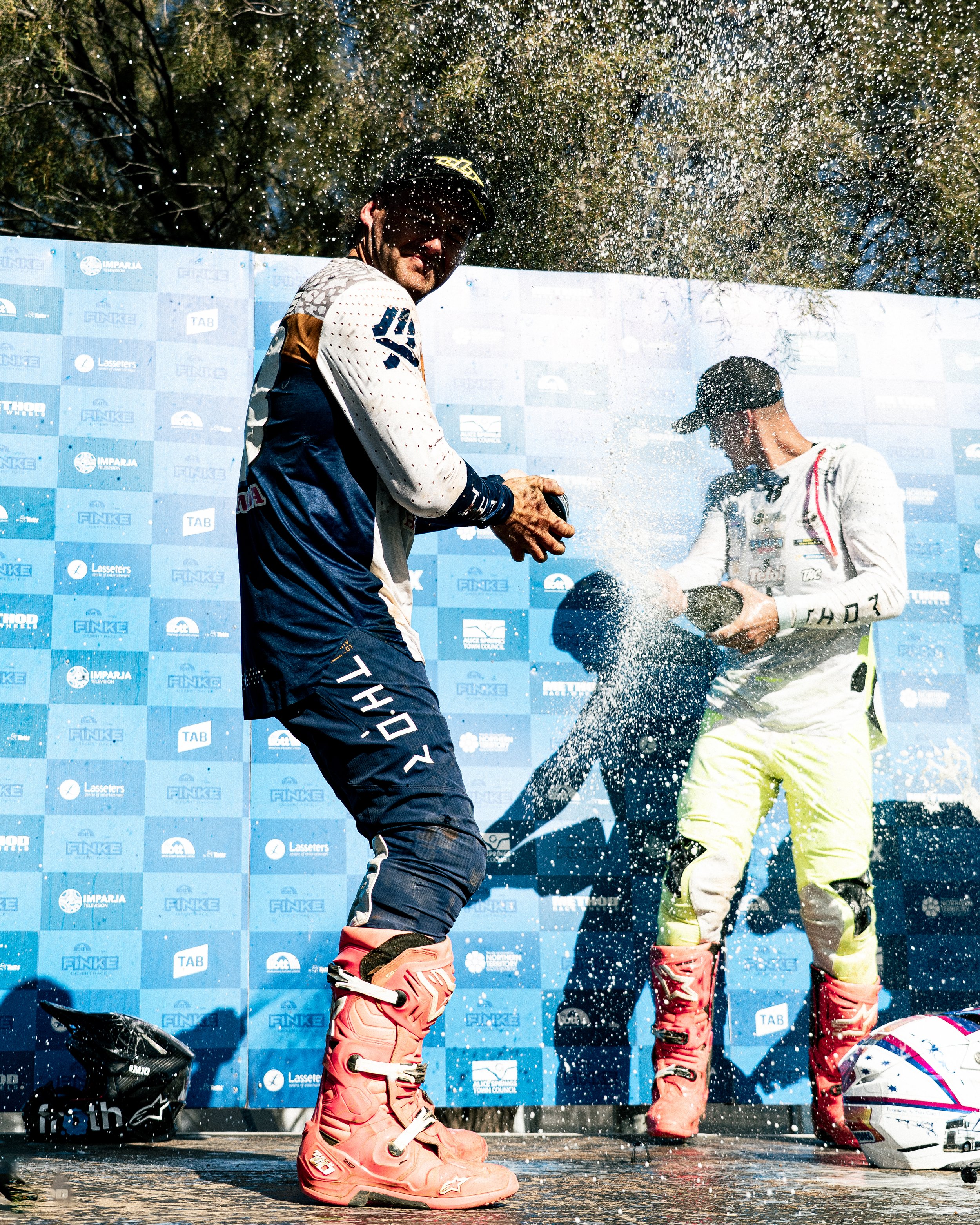Two race car drivers celebrating on a podium, spraying champagne, wearing racing suits and boots, with a blue backdrop featuring various logos.