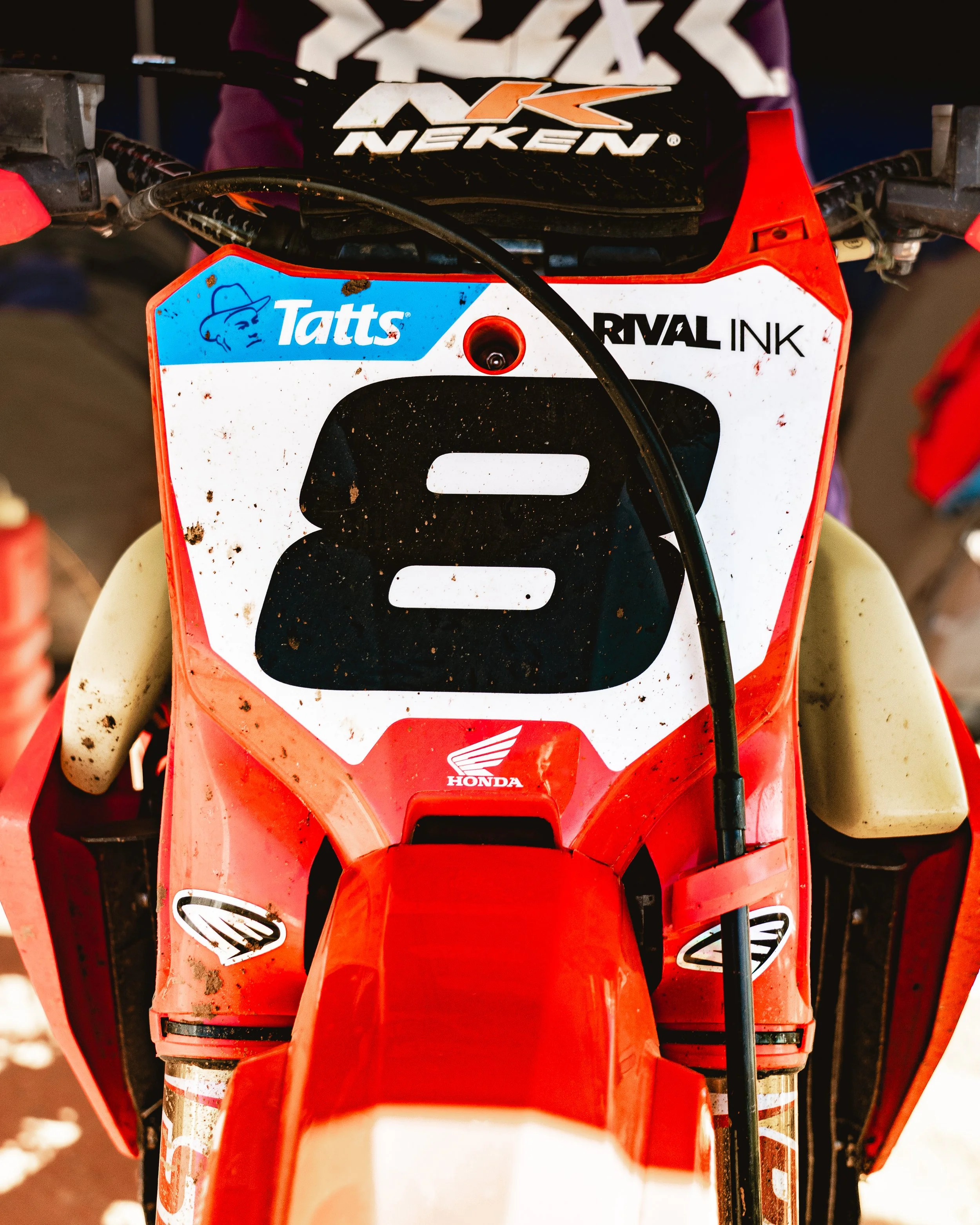 Close-up front view of a dirt bike with a red and white color scheme, featuring various stickers, including 'Tatts', 'Rival Ink', and a Honda logo, with a dirt-covered windshield and handlebars.