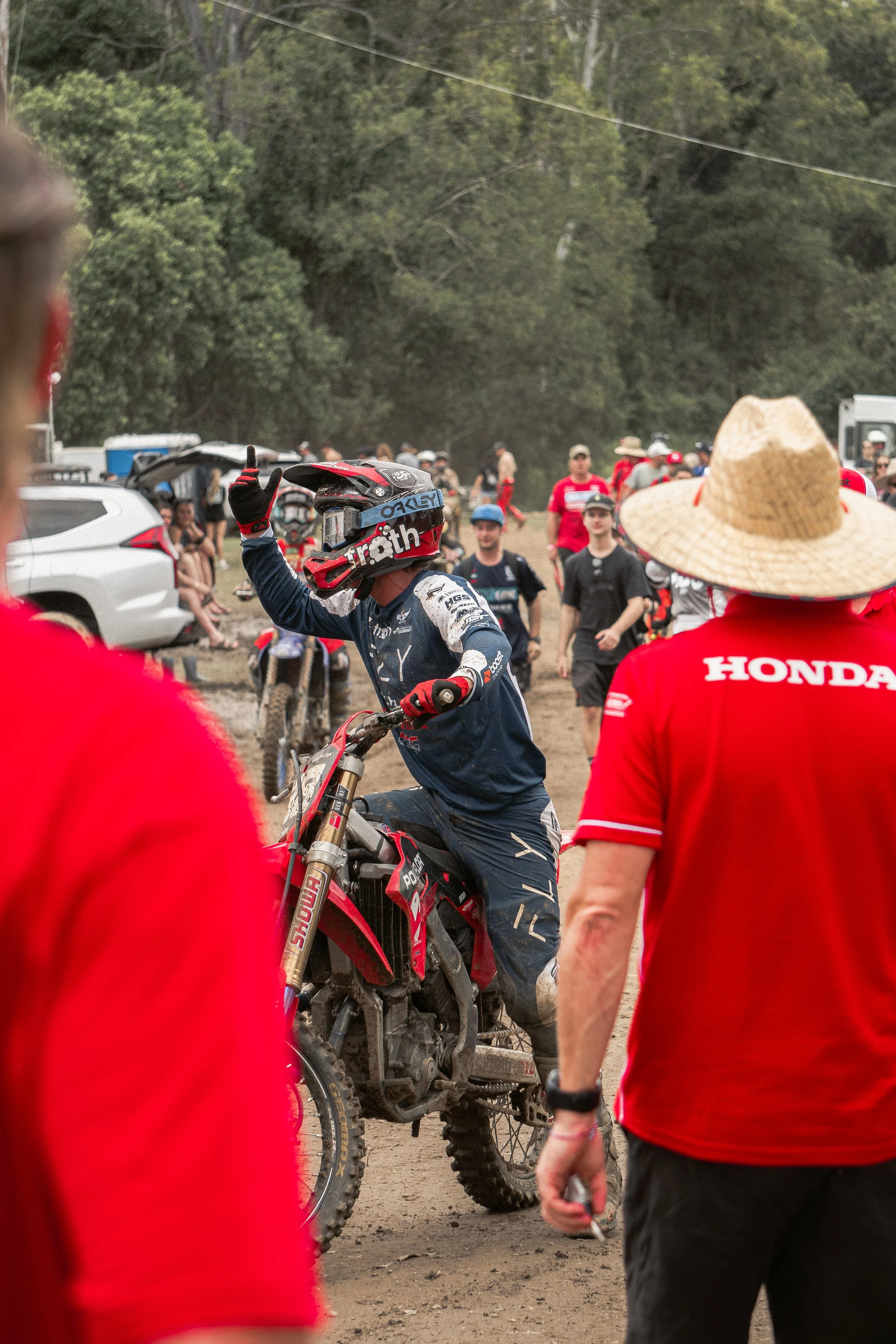 A motocross rider in a blue and white suit and helmet giving a thumbs-up while sitting on a red motorcycle, surrounded by people, some wearing red shirts and straw hats, at an outdoor dirt track.