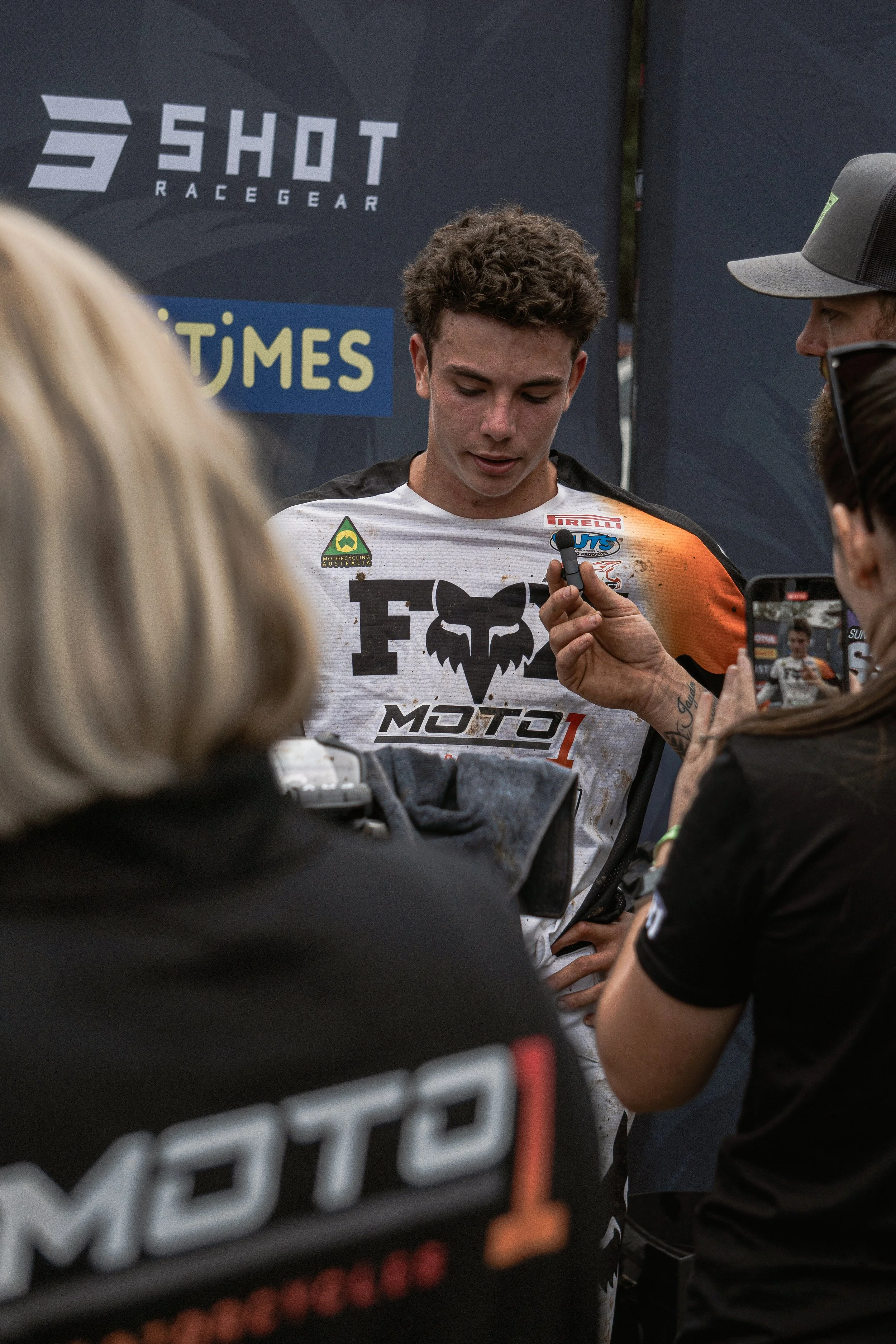 A young male motocross rider is being interviewed by journalists after a race, with a backdrop featuring logos for 5 HIVE Race Gear and other sponsors.