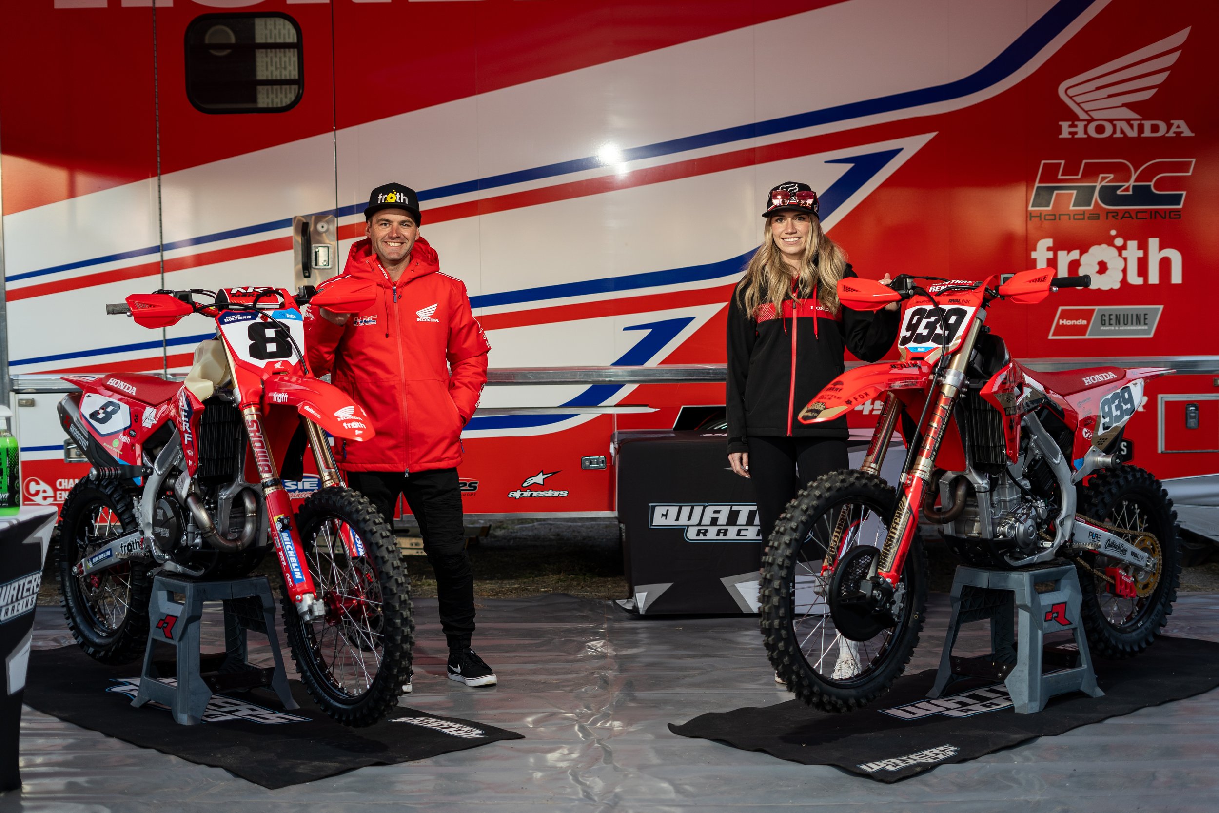 Two motocross riders, a man and a woman, standing next to their red and black motorcycles at a racing event. The man is wearing a red jacket and black cap, and the woman is wearing a black and red jacket and a cap. They are standing in front of a lar