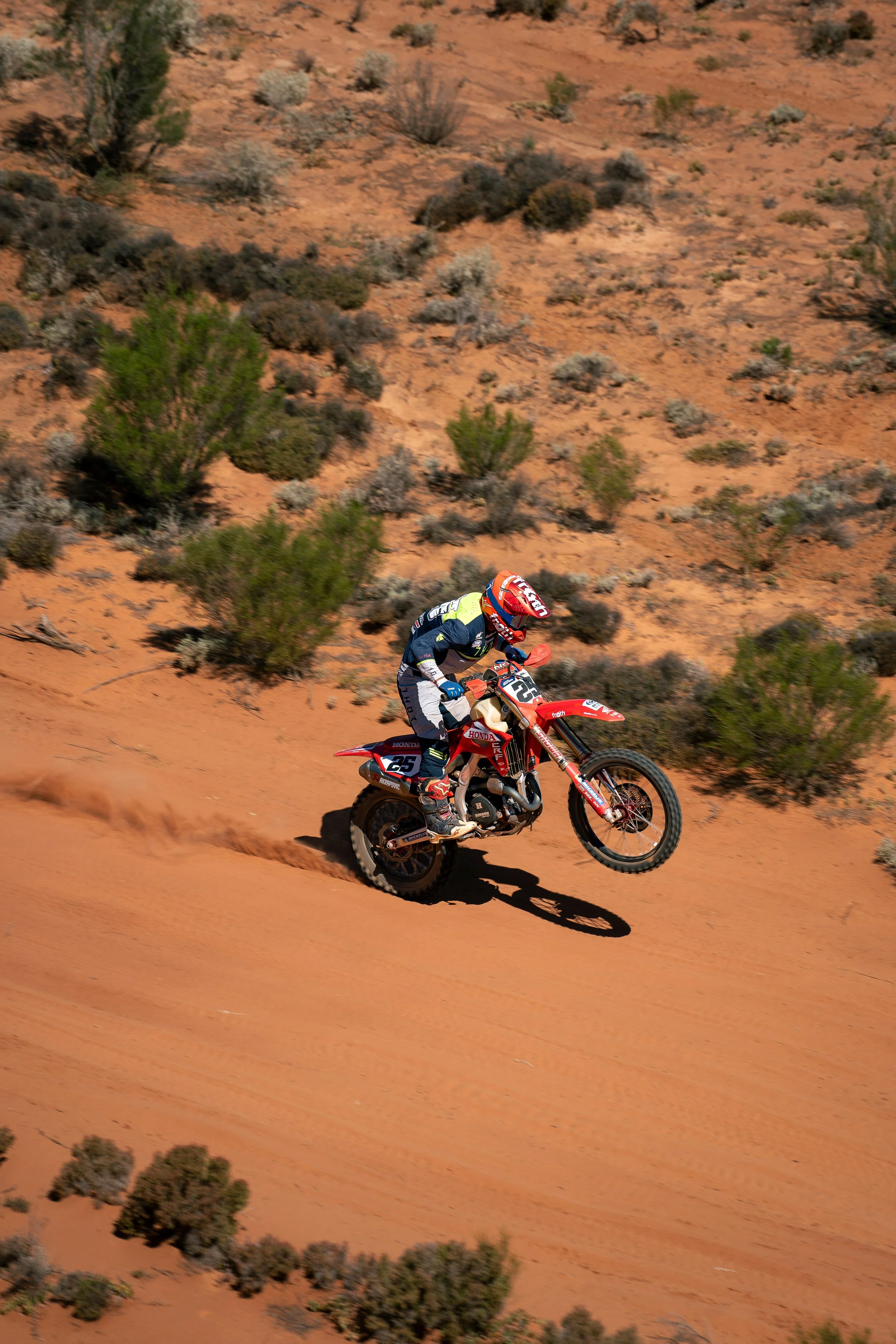 A person riding a dirt bike on a desert trail with bushes and sandy terrain.