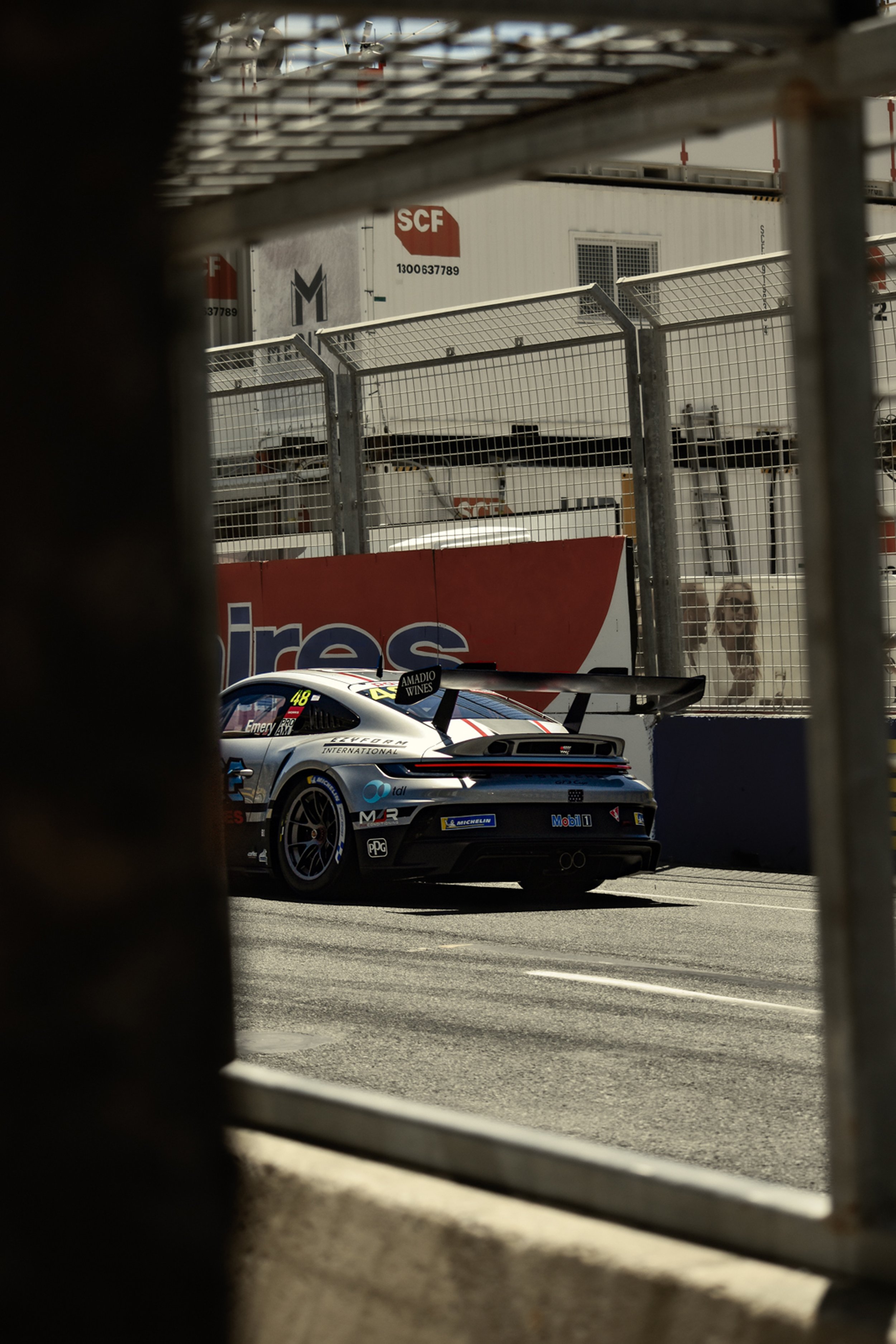 Race car on a racing circuit, viewed through a fence with a background of race track barriers and industrial buildings.