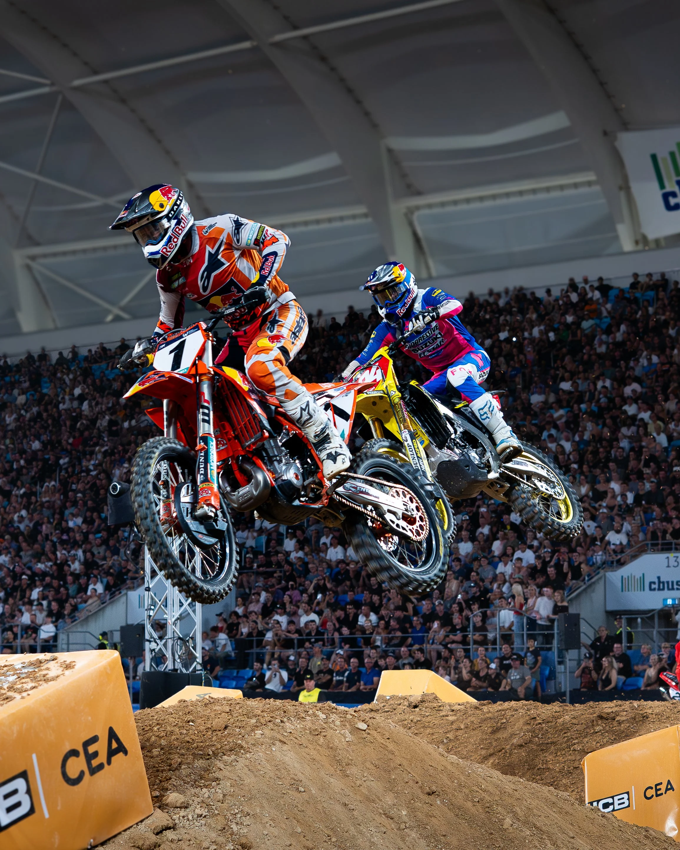 Two motocross racers in mid-air during a race, with a large crowd in the background and dirt jumps below.