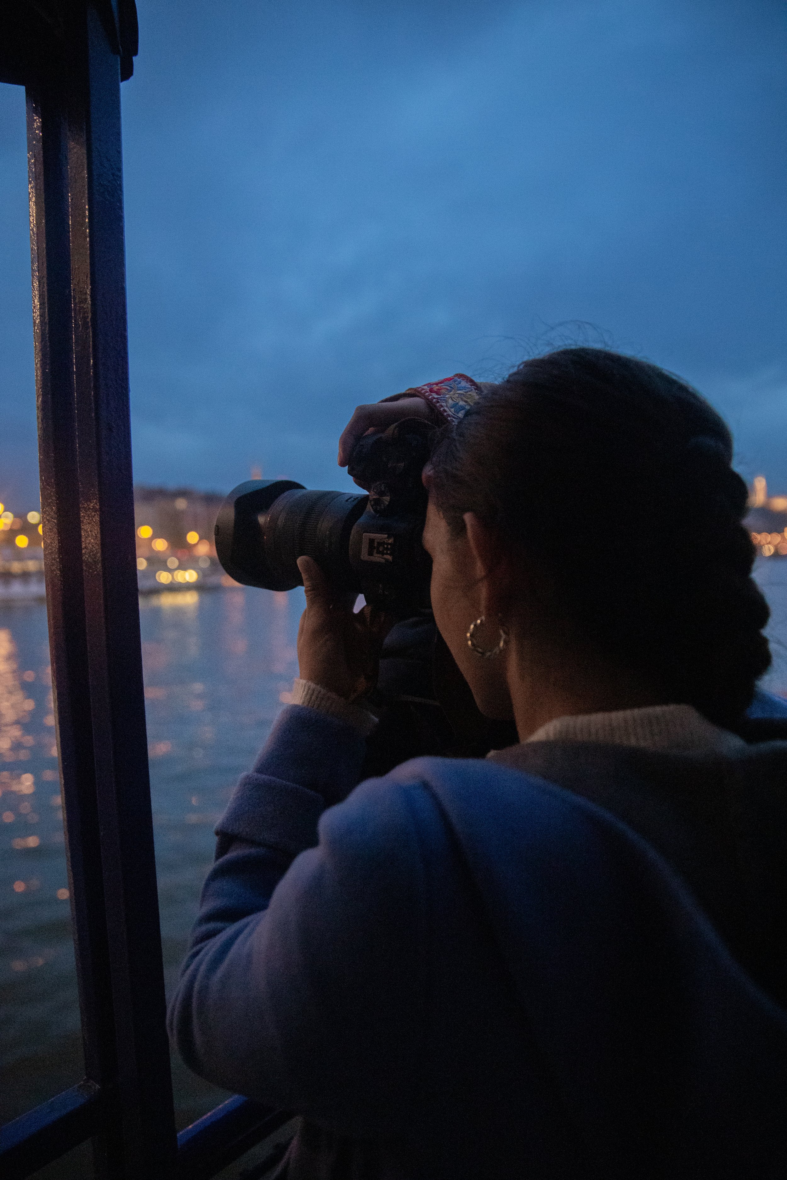 Person with dark hair and earrings taking a photo with a camera on a boat during dusk or night, overlooking a body of water with city lights in the background.