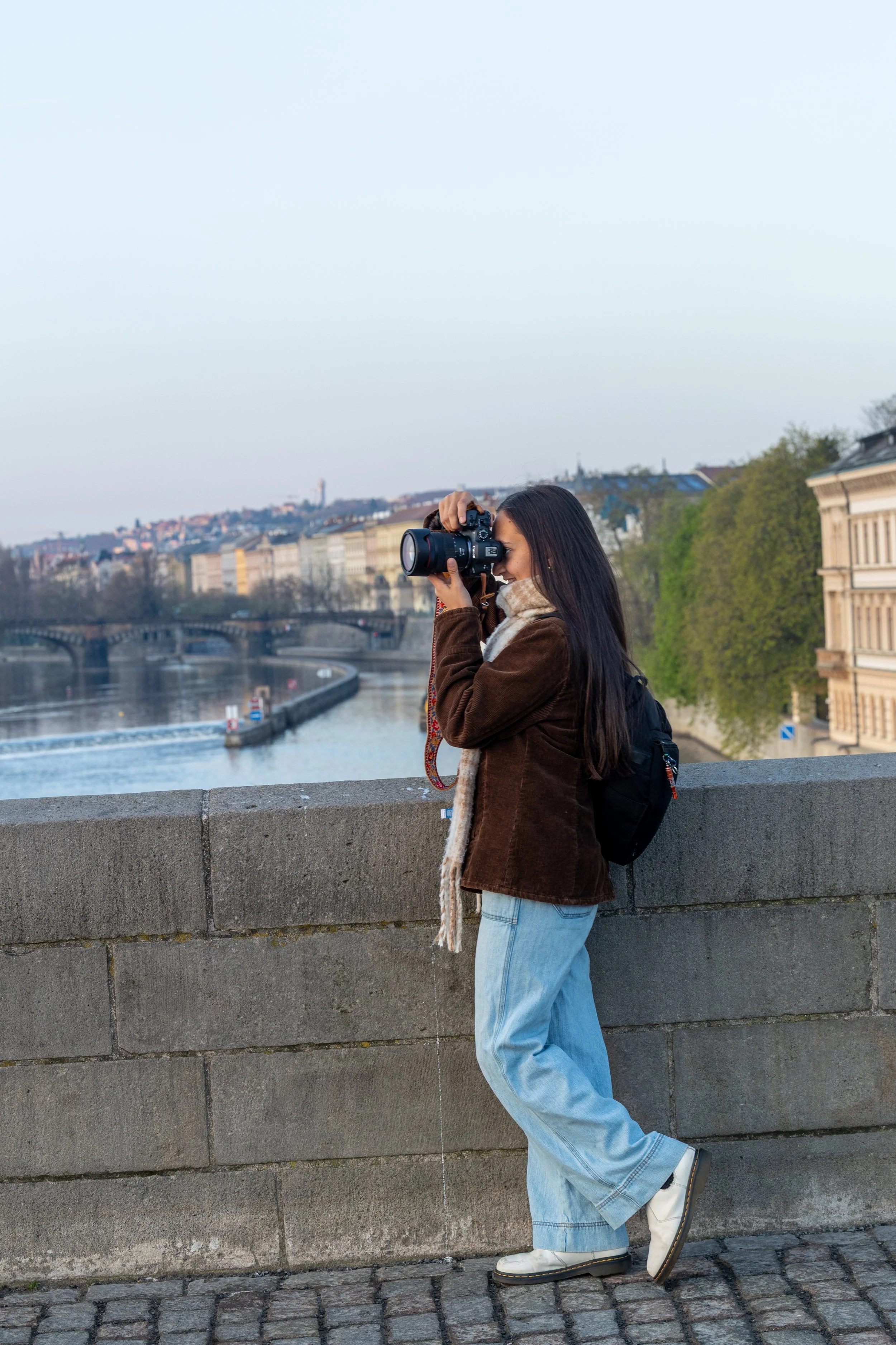 A woman with long dark hair wearing a brown jacket, light blue jeans, and white shoes, taking a photo with a camera on a bridge overlooking a river and cityscape in the background.
