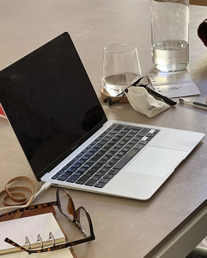 A workspace with a MacBook laptop, glasses of water, a pen, a small pouch, notes, sunglasses, a notebook, and wristbands on a table.