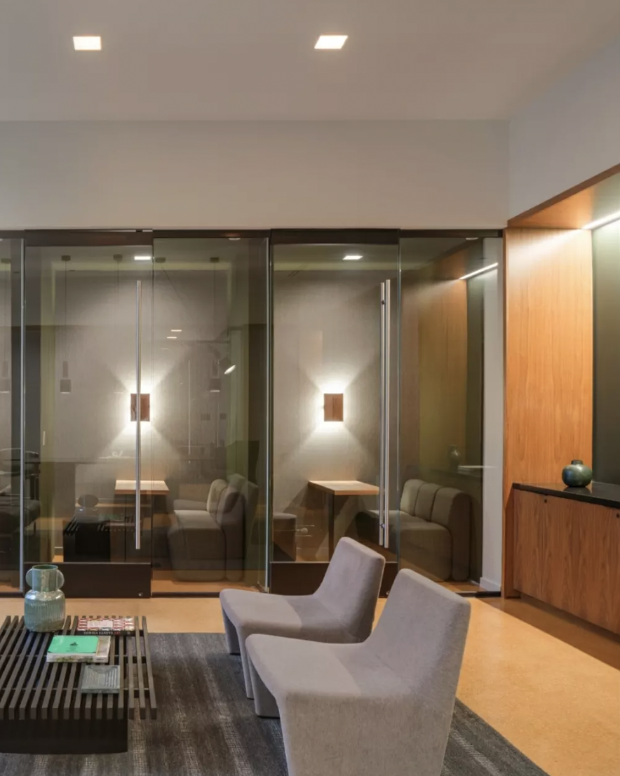 Modern hotel lobby with two light gray chairs, a wooden coffee table with magazines and vases, a dark rug, a wooden wall with decorative vase, and glass doors leading to a private seating area with wall-mounted lights.