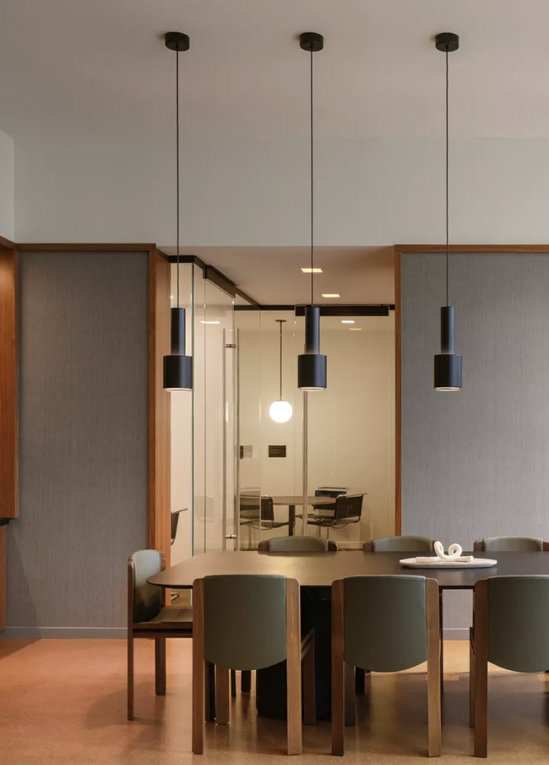 Modern dining room with a wooden table and six green chairs, three black pendant lights hanging from the ceiling, and a glass-walled conference room in the background.