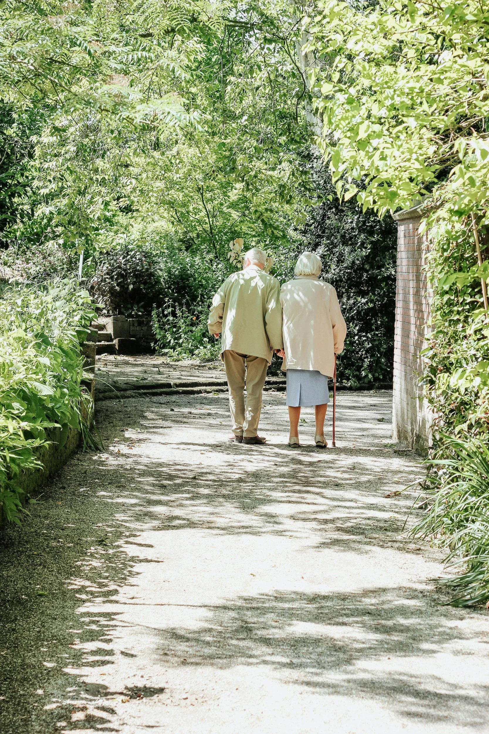 An elderly couple walks down a shaded garden path, surrounded by lush green trees and plants. The man is wearing a beige jacket and trousers, while the woman wears a light pink coat, a gray skirt, and uses a walking cane.