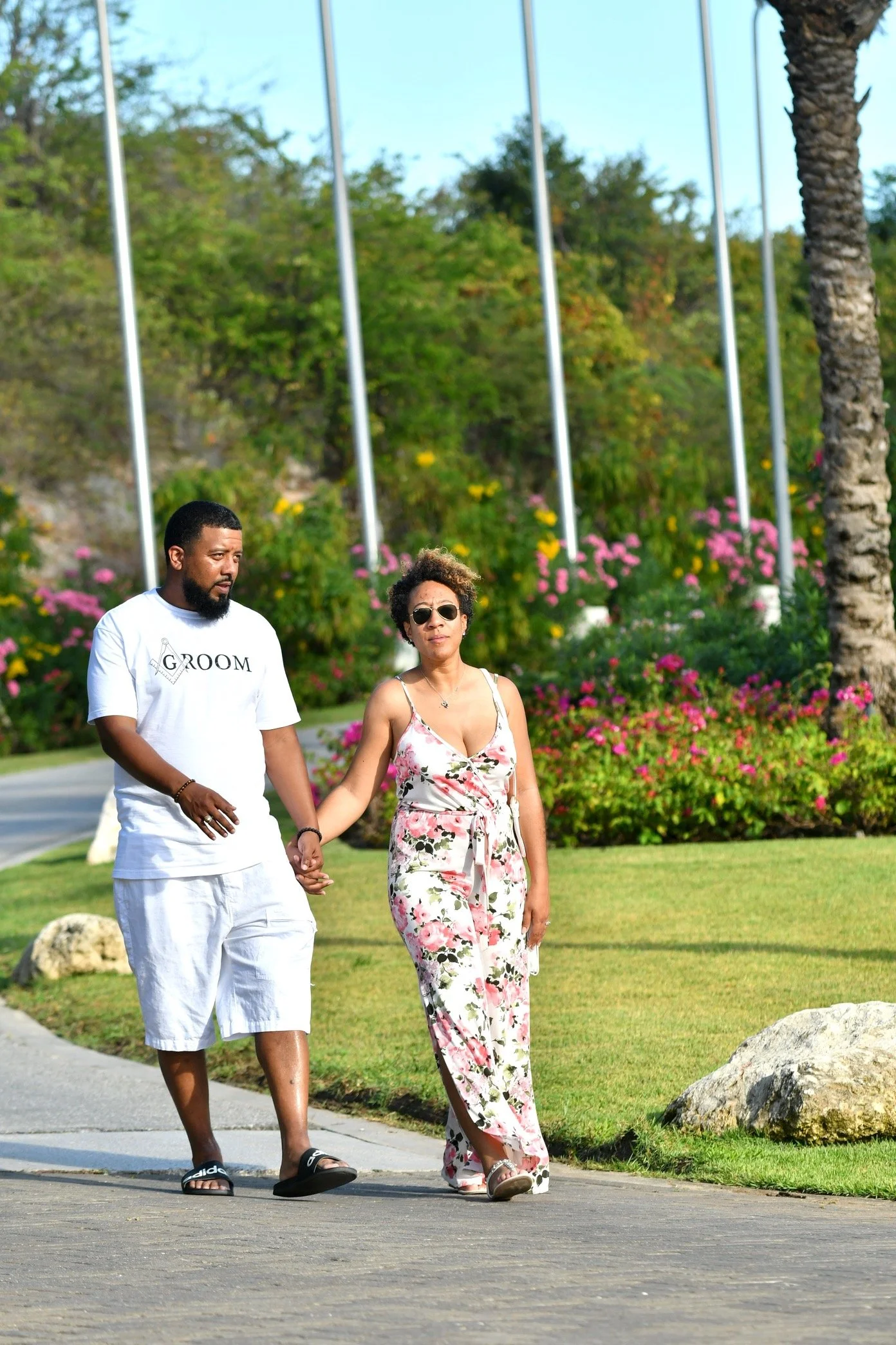A man and woman walking hand in hand outdoors near a garden with pink flowers. The man wears a white t-shirt with 'GROOM' printed on it and white shorts. The woman wears sunglasses and a floral jumpsuit.