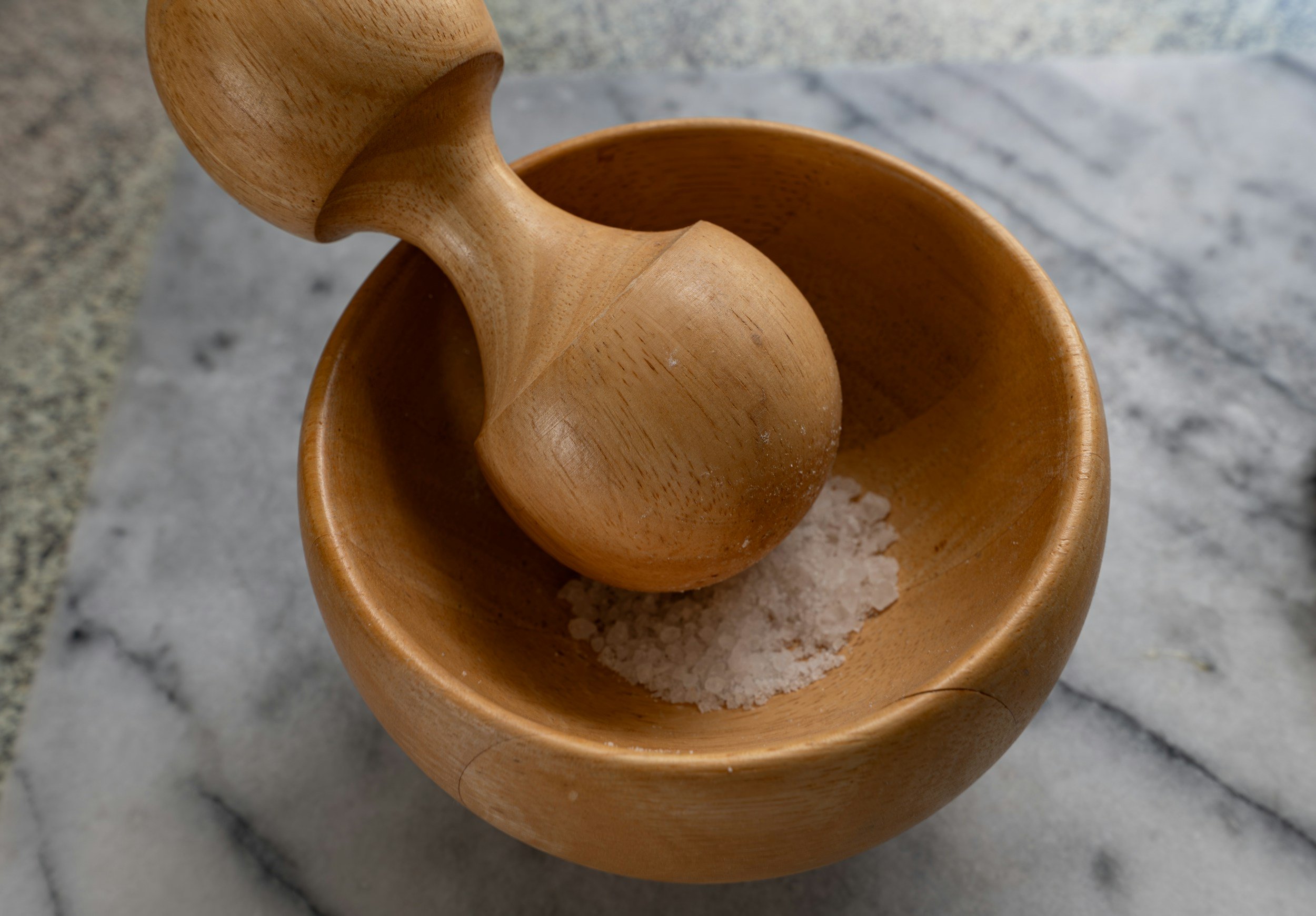 A wooden mortar and pestle with salt inside, resting on a marble surface.