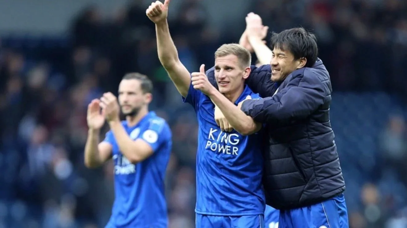 Leicester City soccer players celebrating on the field, one is giving a thumbs up, another is hugging him, all wearing blue jerseys with 'King Power' logo.