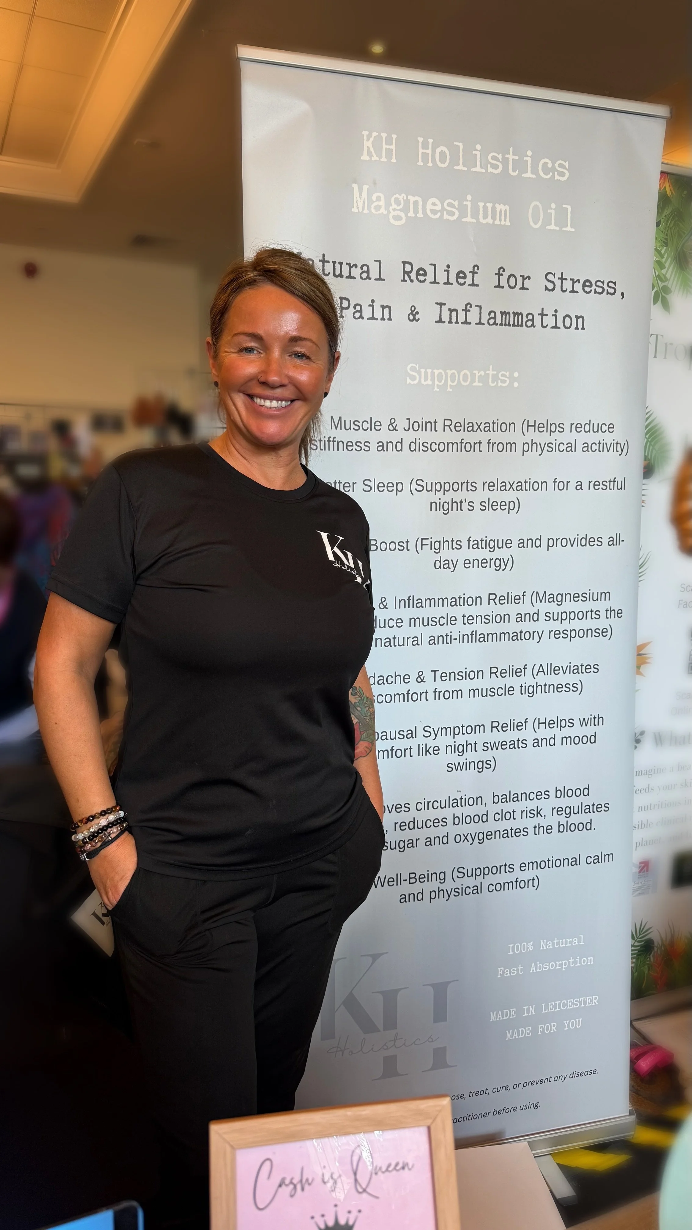A smiling woman standing in front of a display banner for KH Holistics Magnesium Oil, which lists its natural relief benefits for stress, pain, and inflammation, along with various support benefits. She is wearing a black KH Holistics t-shirt and black pants, with bracelets on her left wrist.