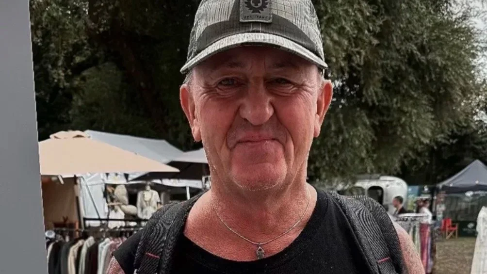 DJ Danny Rampling is wearing a cap, black sleeveless shirt, and a small necklace is smiling slightly at an outdoor market with tents and trees in the background. He is holding a bag with magnesium oil in from KH Holistics.