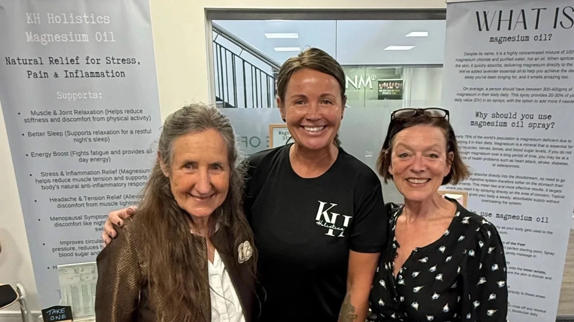Three women smiling and posing in front of informational posters about magnesium oil at an indoor event.