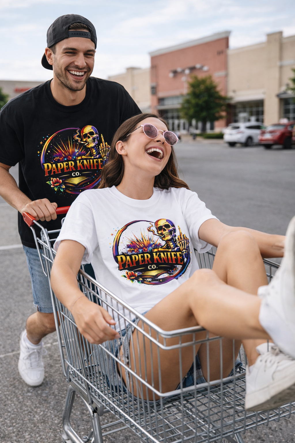 A young woman sitting in a shopping cart with a big smile, wearing sunglasses and a t-shirt with a colorful skull and floral design, being pushed by a young man with a black cap and a matching t-shirt, both in a parking lot.