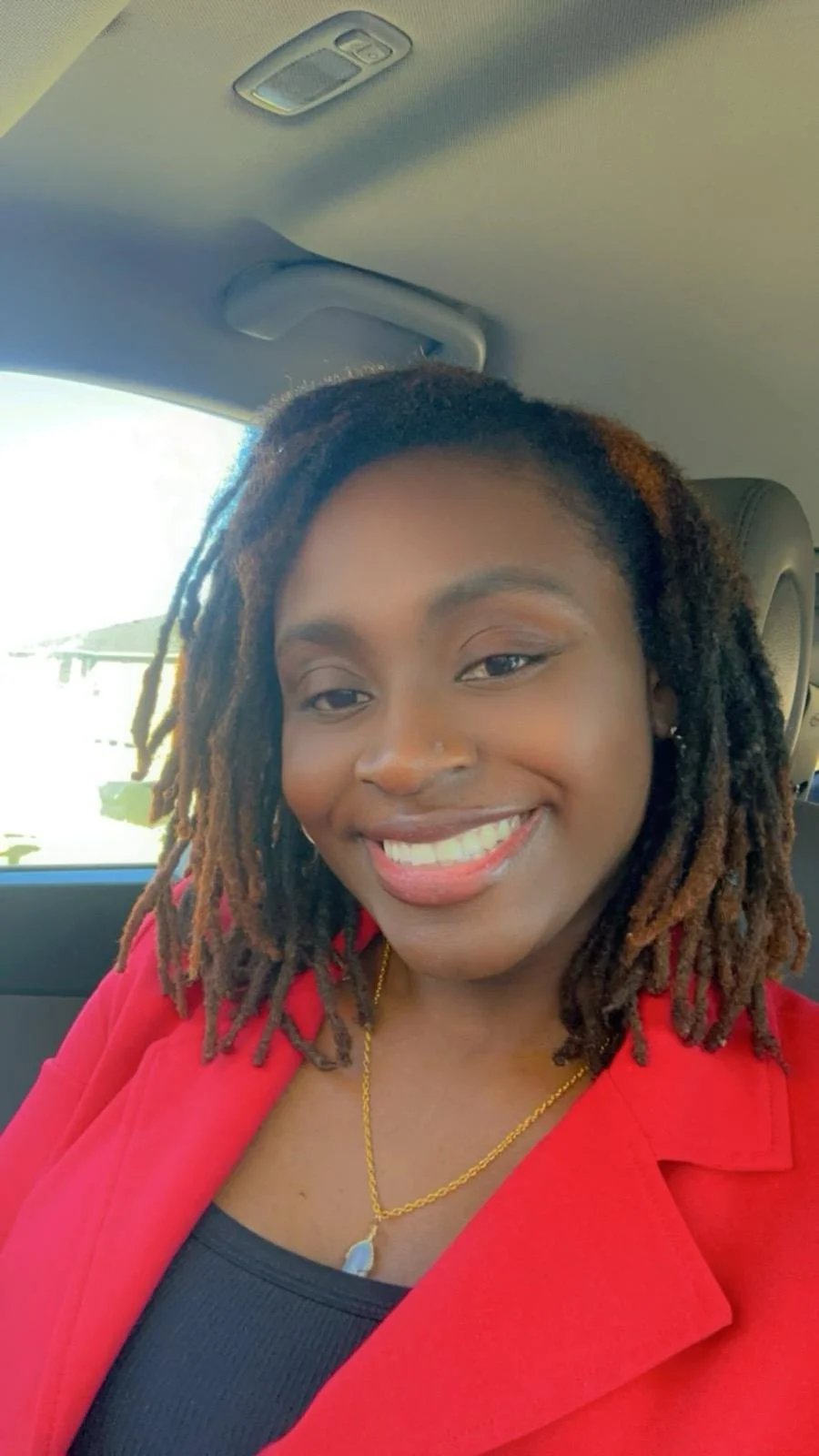 A woman with dreadlocks smiling, wearing a red blazer and a gold necklace, sitting in a vehicle.