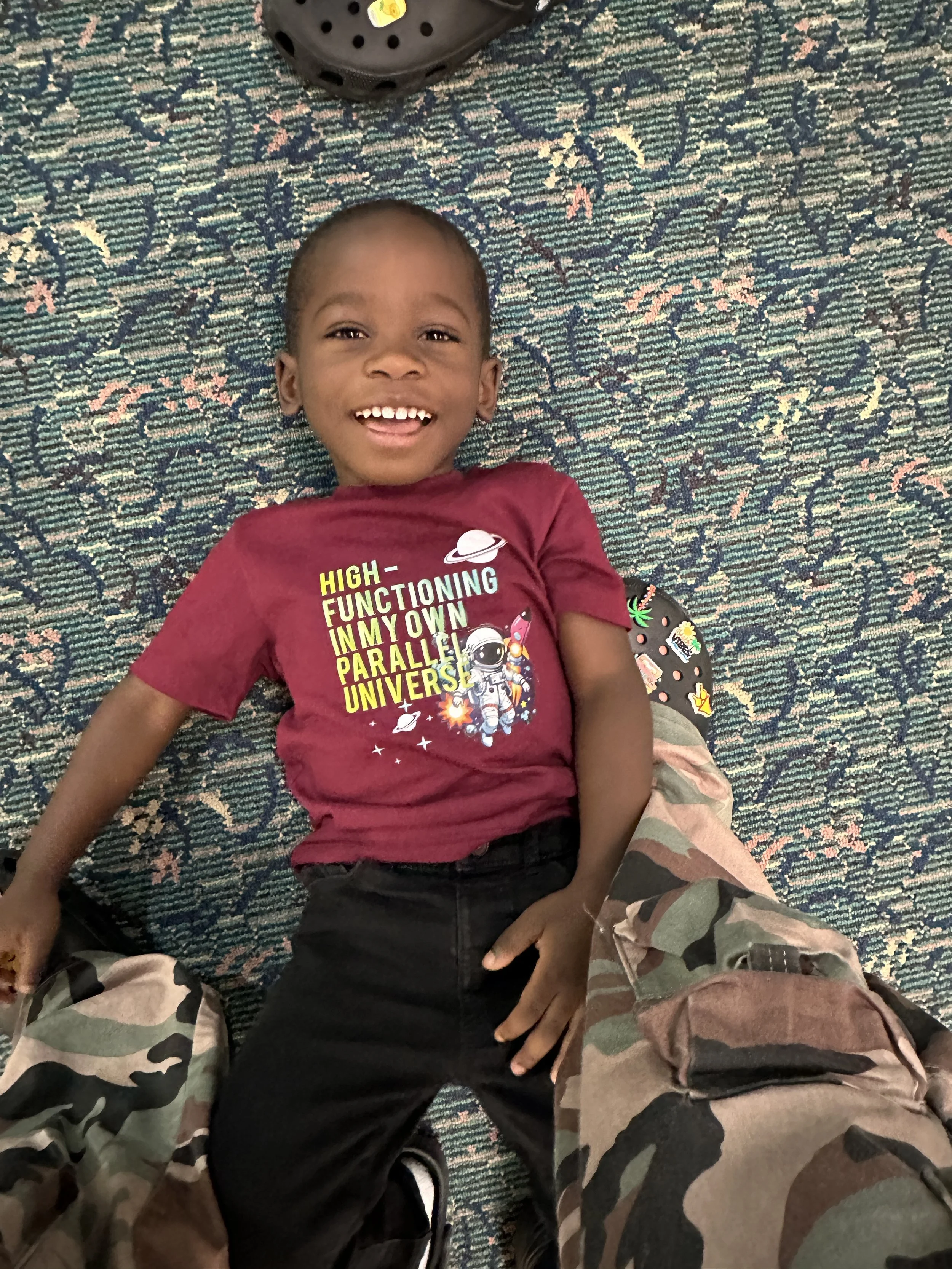 A young boy lying on a patterned carpet, smiling, wearing a maroon T-shirt with space-themed graphics, and black pants.