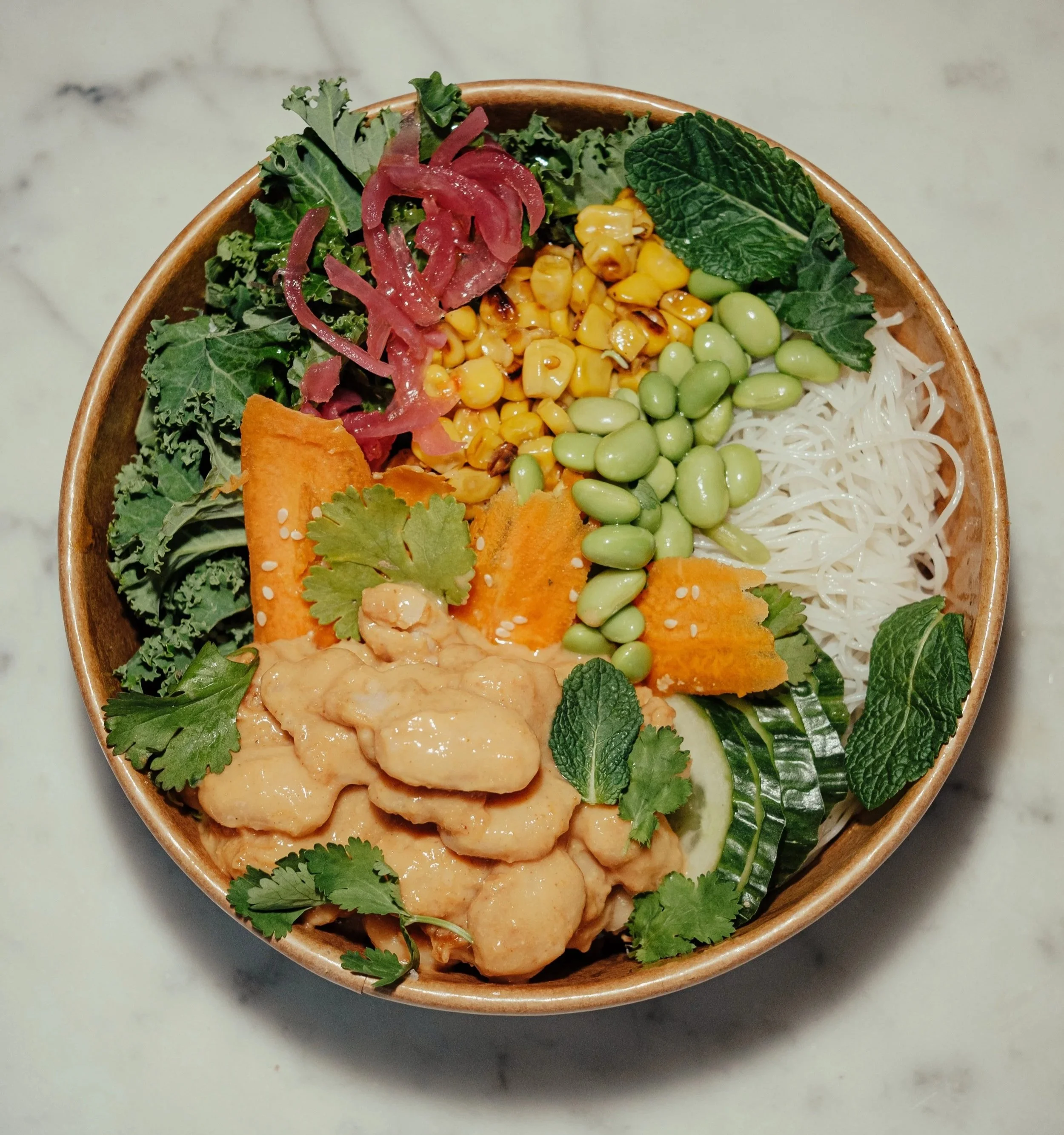 Greens & Grains salad bowl. Close-up of a bowl filled with various fresh vegetables and herbs, including kale, mint, edamame, corn, shredded noodles, and slices of sweet potato, garnished with cilantro leaves on a marble surface.