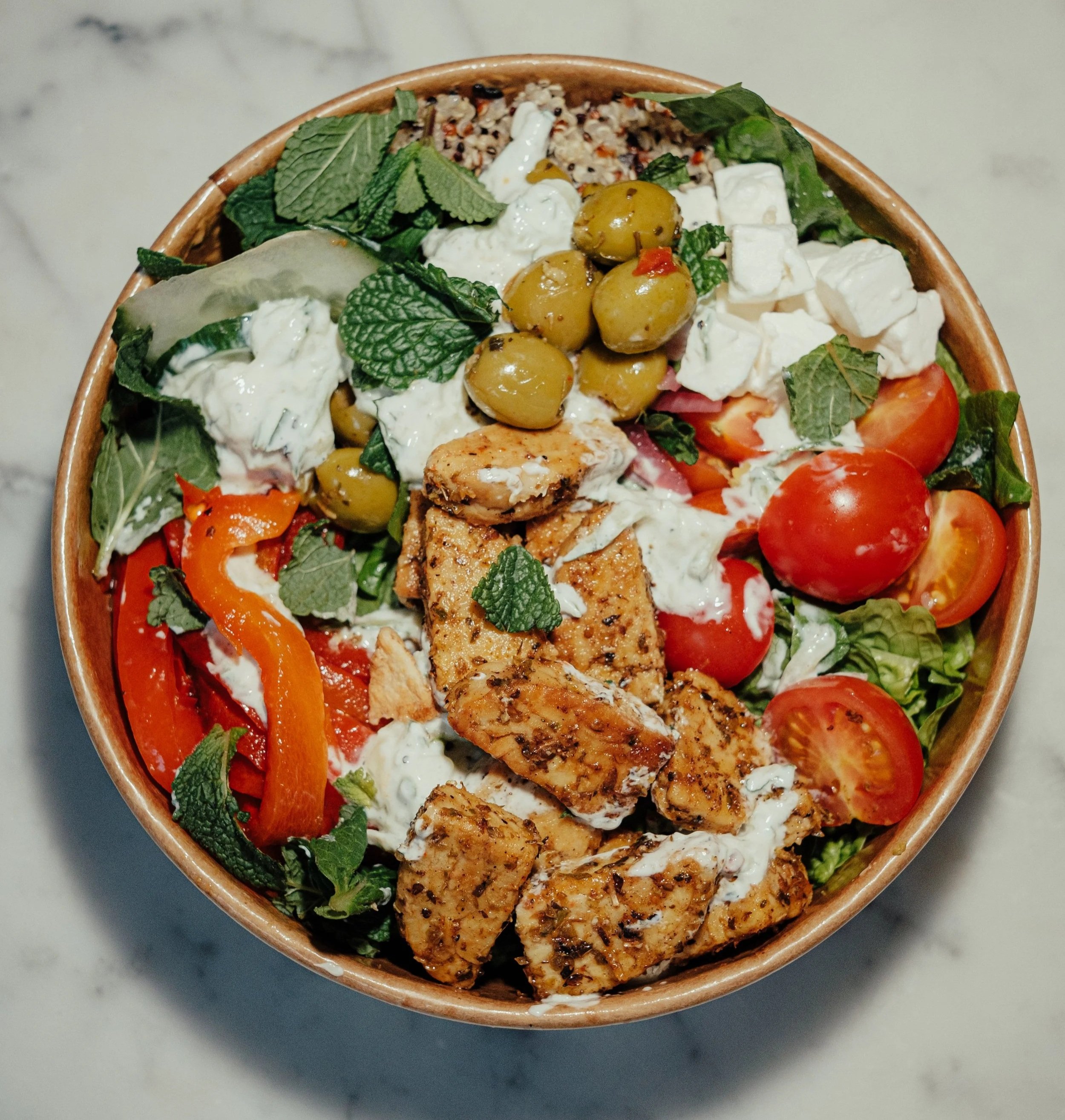 Greens & Grains salad bowl. A bowl of salad with cherry tomatoes, grilled chicken, green olives, feta cheese, cherry tomatoes, and fresh mint leaves on top, set on a white marble surface.