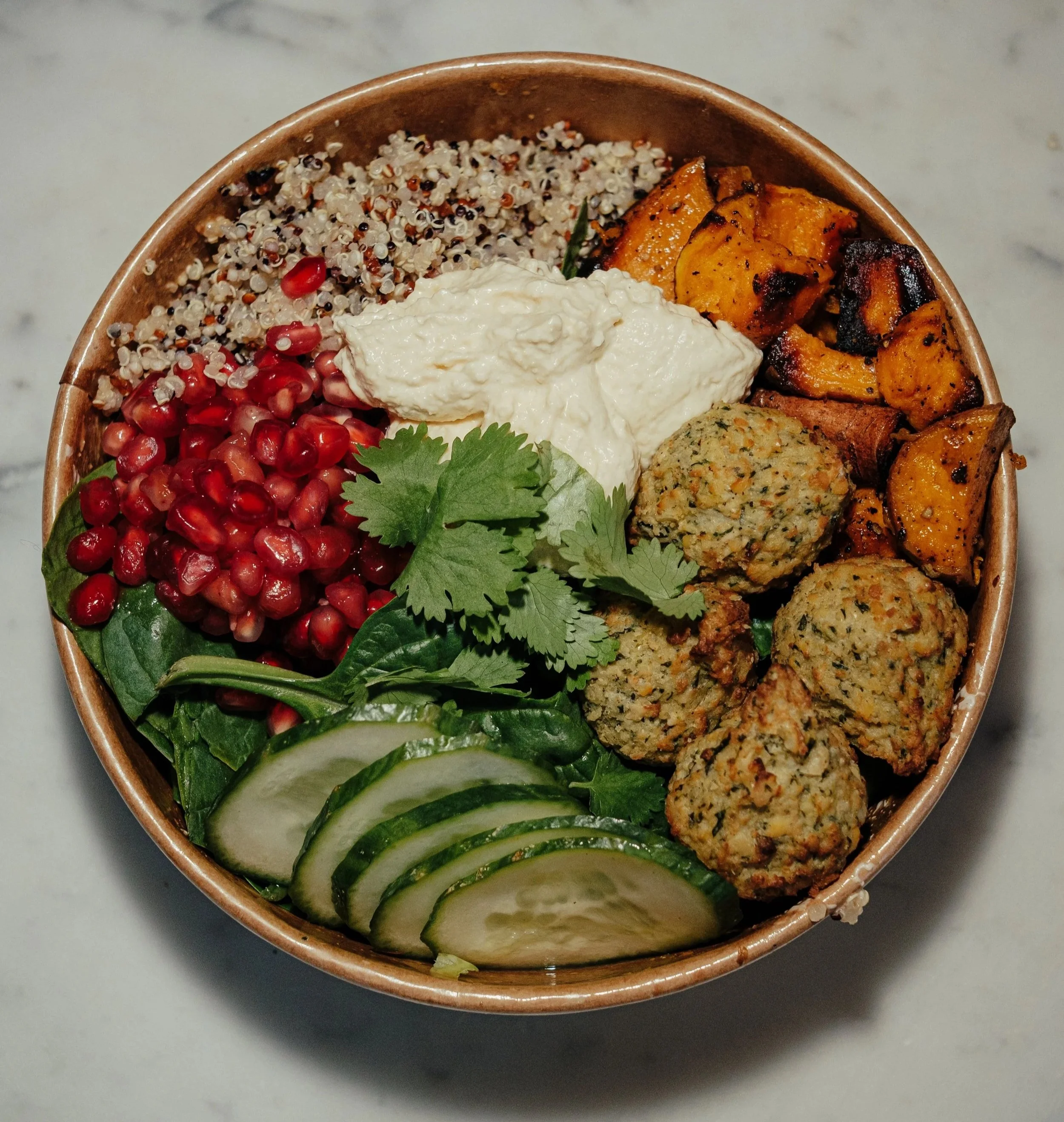 Greens & Grains salad bowl. A bowl containing pomegranate seeds, cucumber slices, a dollop of yogurt, roasted sweet potatoes, and falafel served on a bed of lettuce and cilantro.